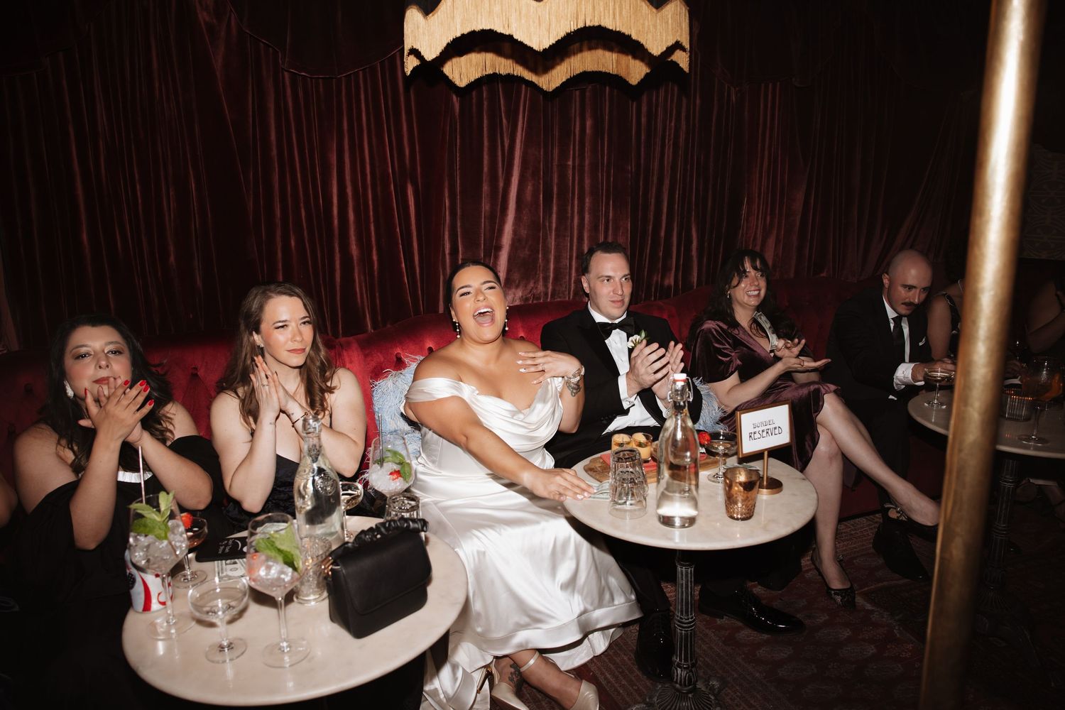 Bride in white dress celebrates with wedding guests at a glamorous dark red velvet reception venue.