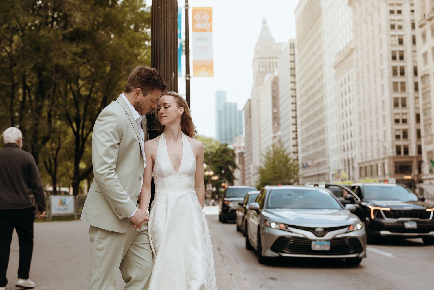 Bride and groom walk hand in hand on a Chicago city street, bride in white gown, groom in tan suit.