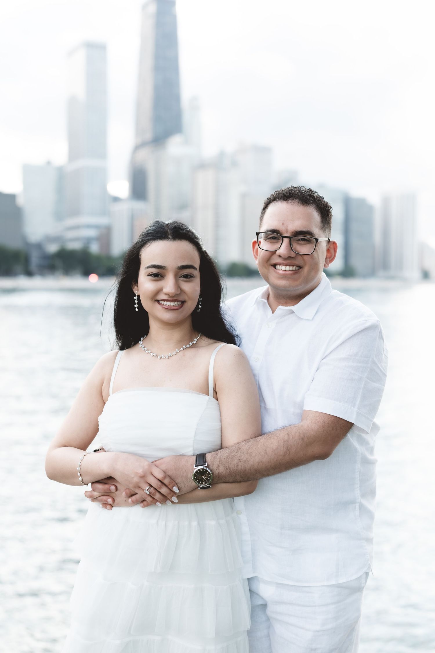Smiling couple in white attire embracing with Chicago skyline and lake in background.