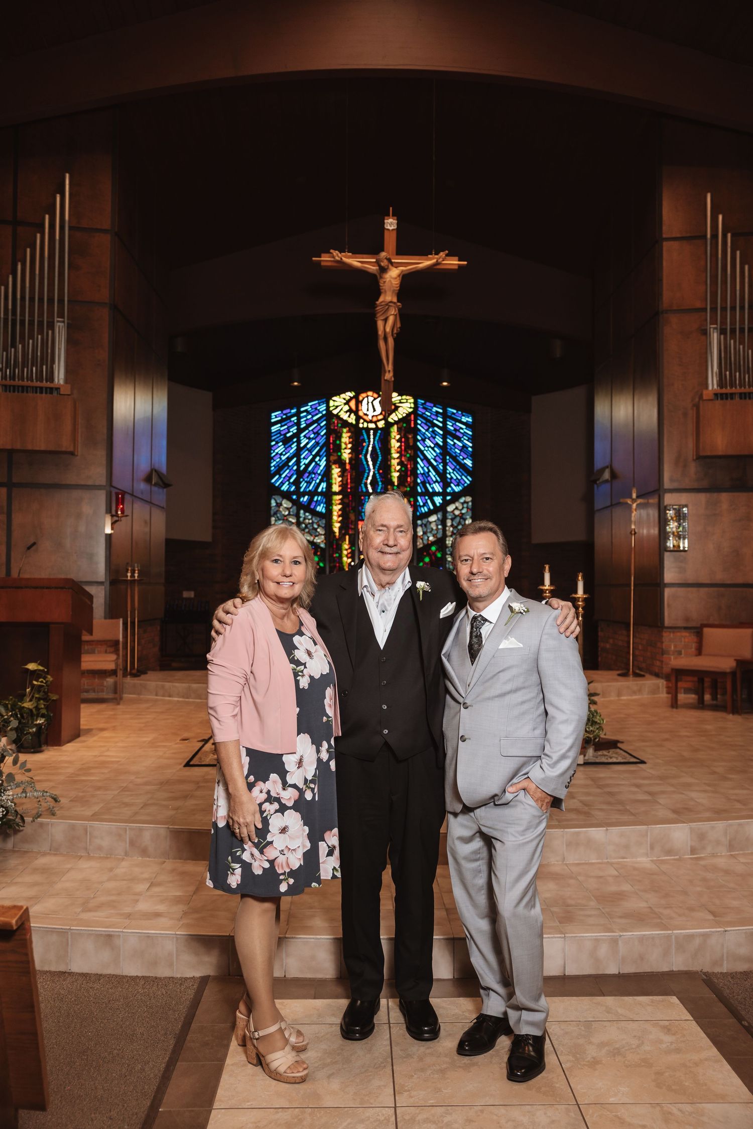 Groom in gray suit poses with older couple in front of church altar with crucifix and stained glass.
