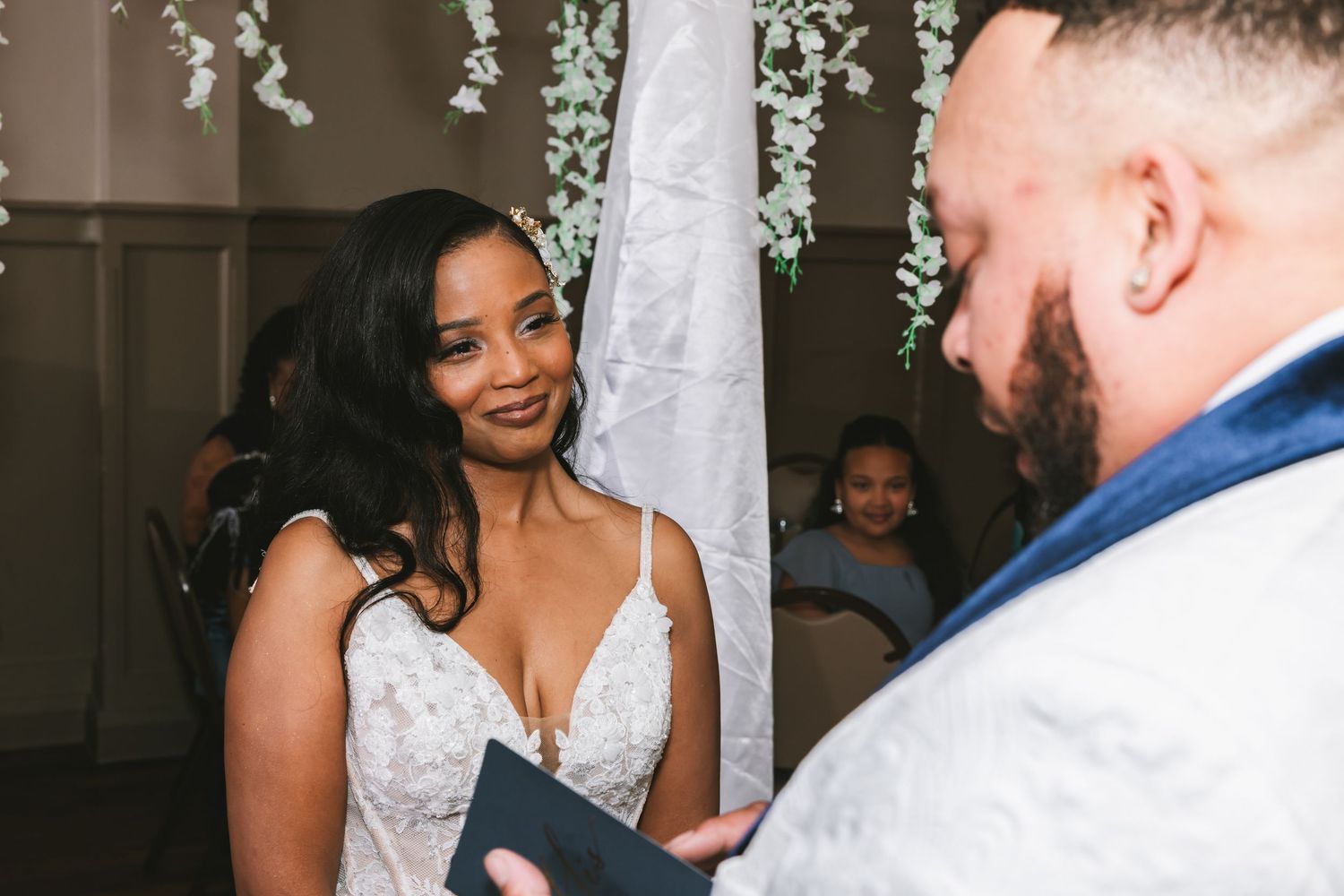 Bride in lace gown smiling lovingly at groom while exchanging vows beneath white floral wedding arch.