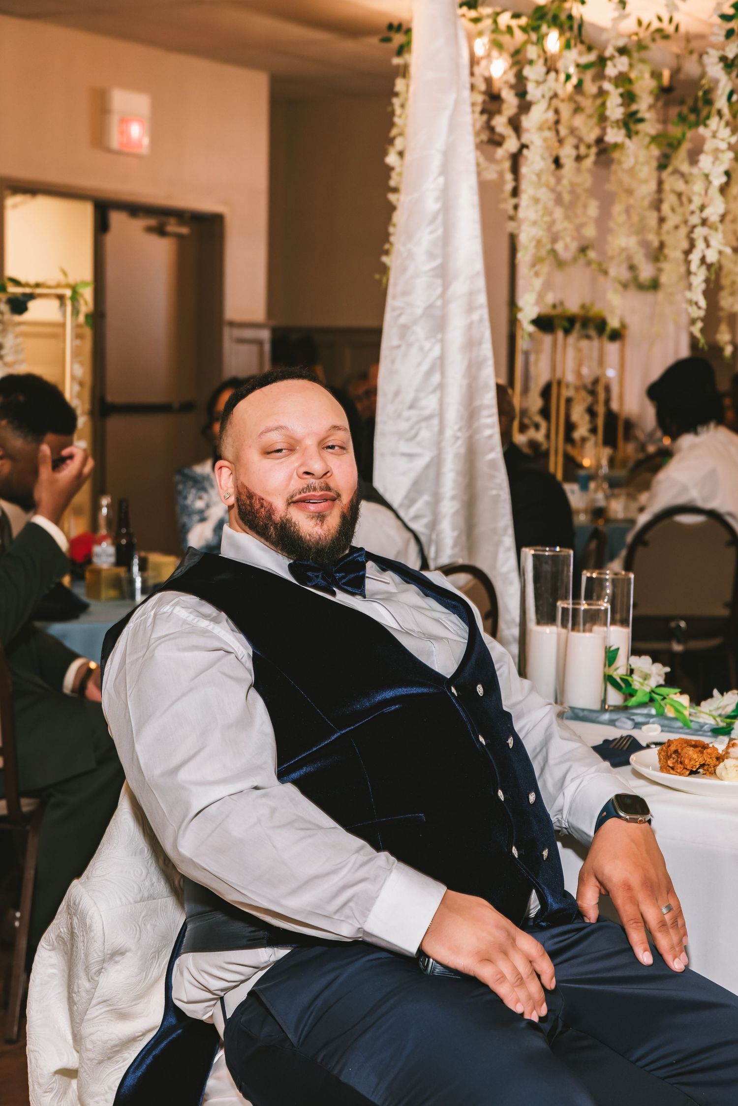 A bald man with a beard wearing a navy vest and bow tie relaxes at a wedding reception table.