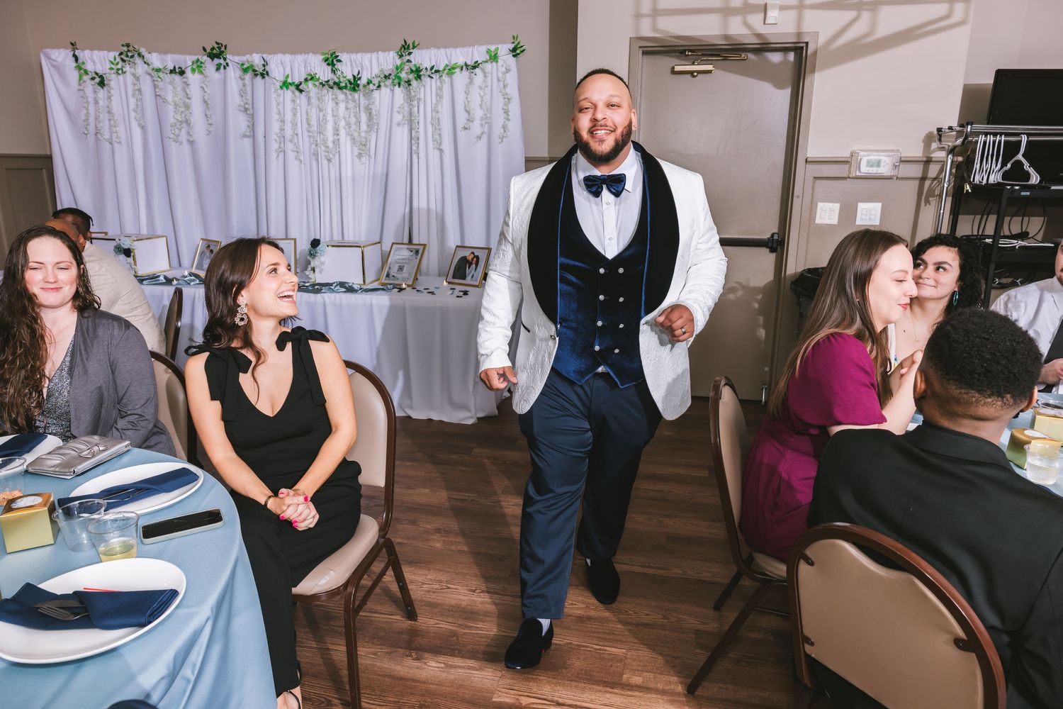 Groom in white tuxedo walks between tables as wedding guests look on at reception venue.