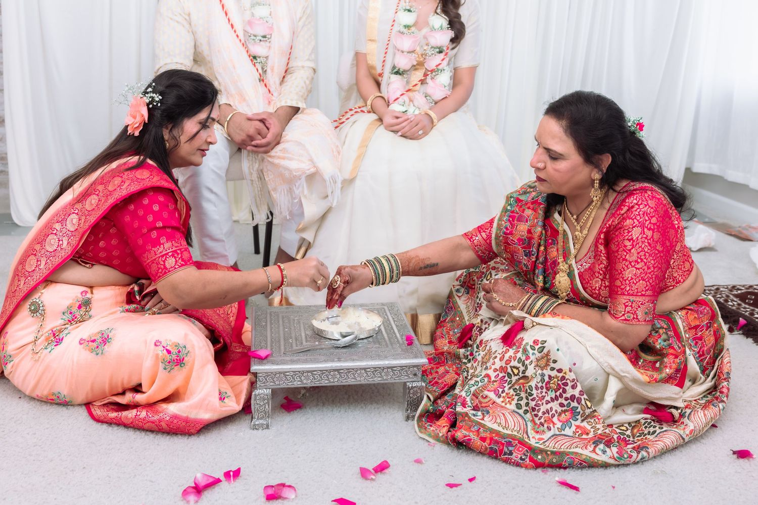 Two women in vibrant red and pink sarees perform a traditional Indian wedding ritual seated on the floor with rose petals.
