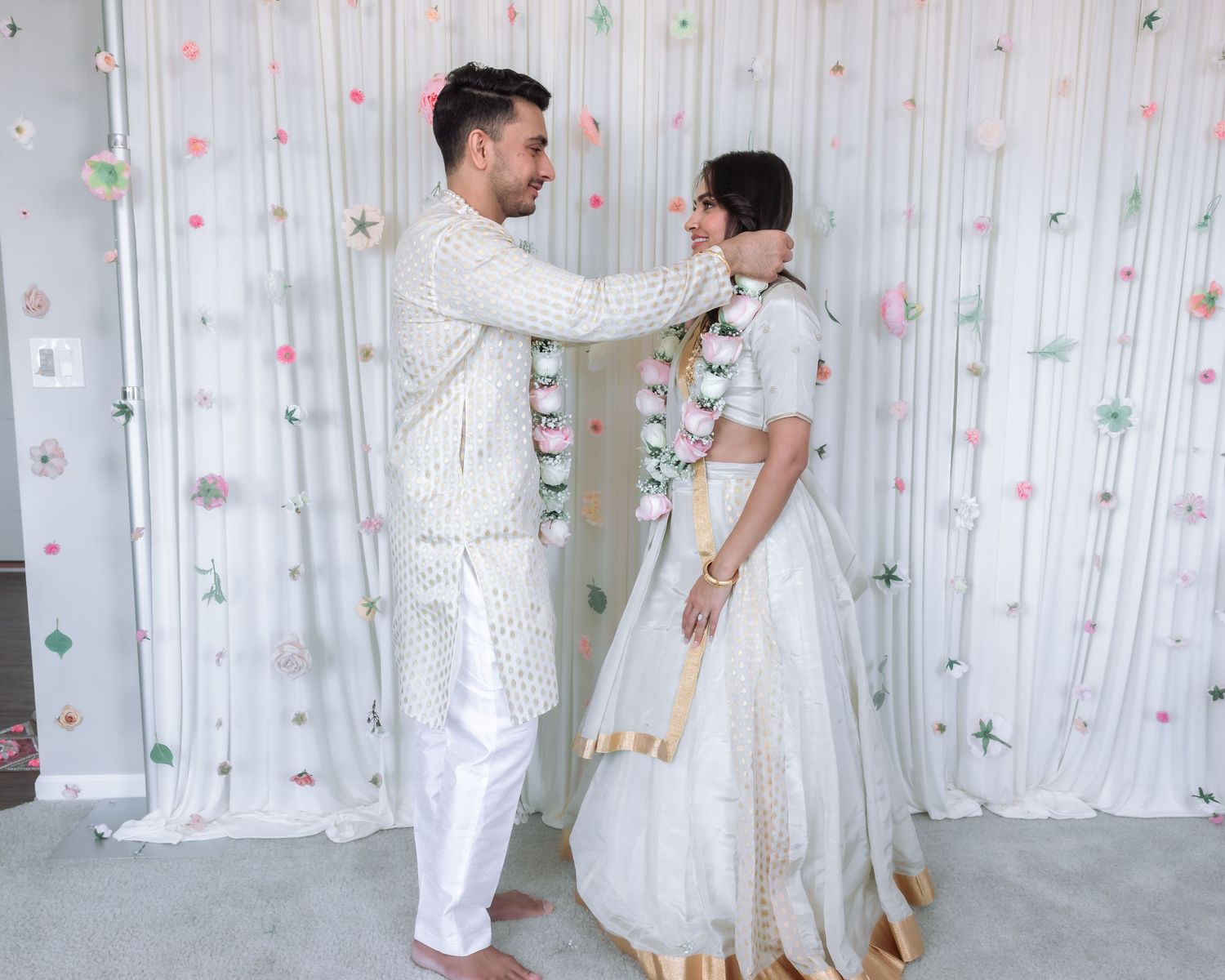 Indian groom places floral garland on bride in white lehenga during traditional engagement ceremony.