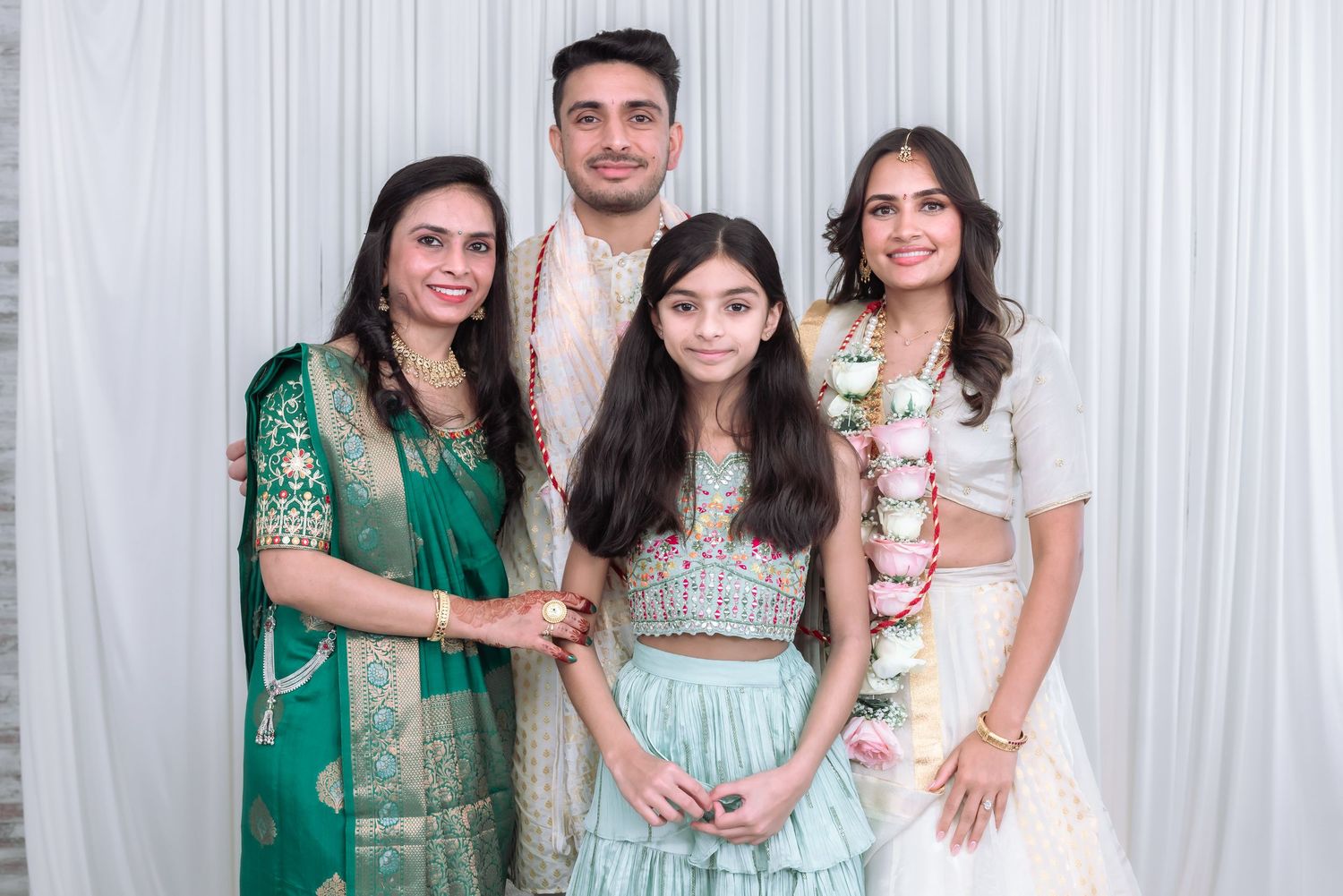 Indian wedding couple in white attire with garlands poses with two women in teal and green traditional Indian clothing.