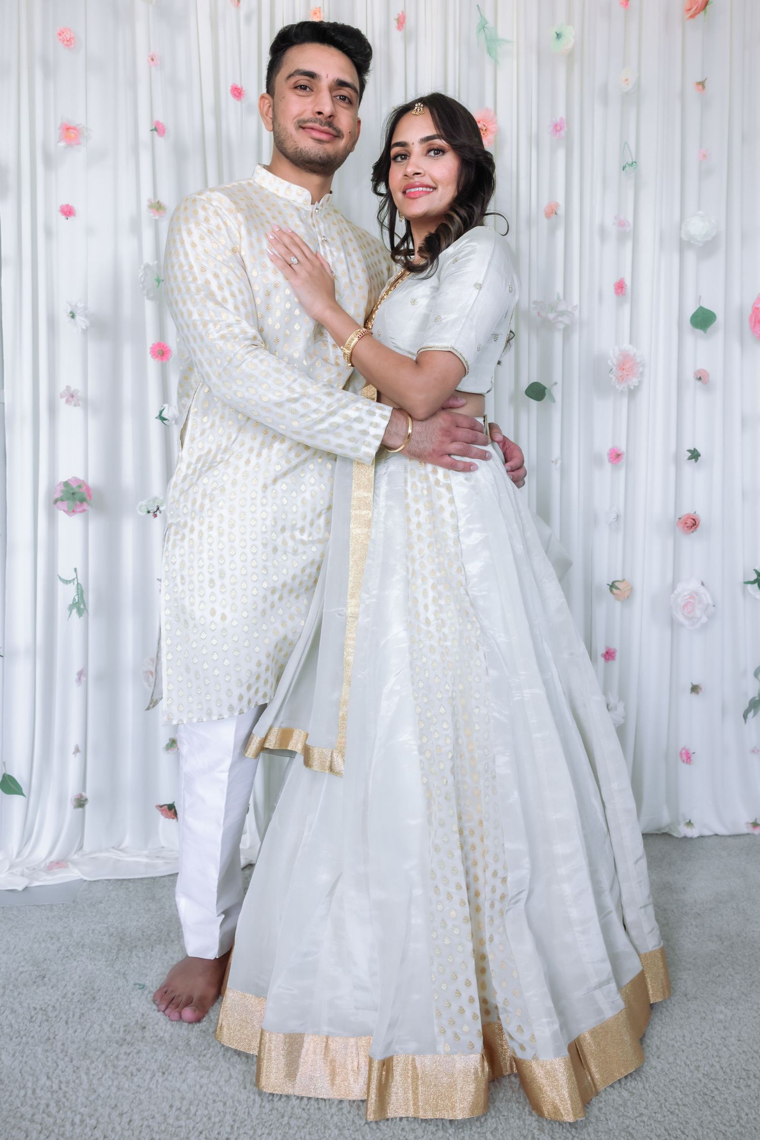 South Asian couple embracing in matching white and gold traditional wedding attire before floral backdrop.