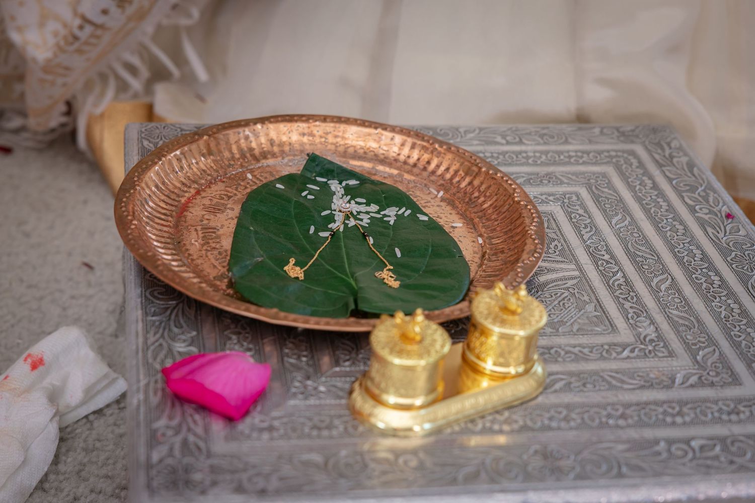 Traditional Indian puja thali with betel leaves, brass kalash vessels, and pink kumkum on silver table.