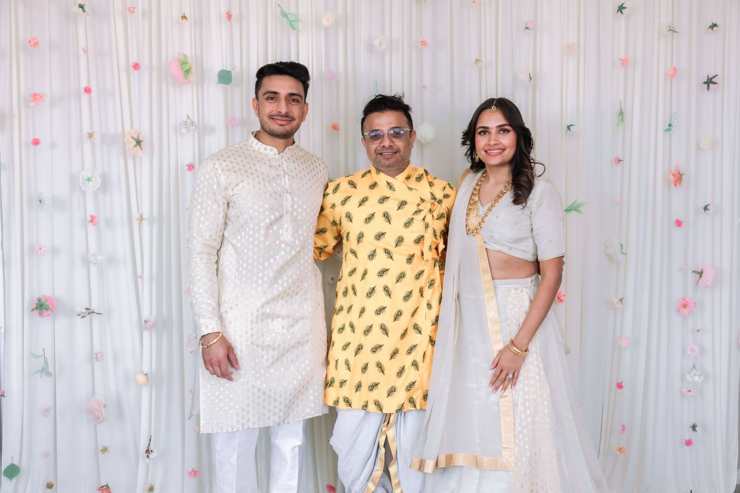 Three young adults in elegant Indian traditional wear pose together at festive celebration with floral backdrop.