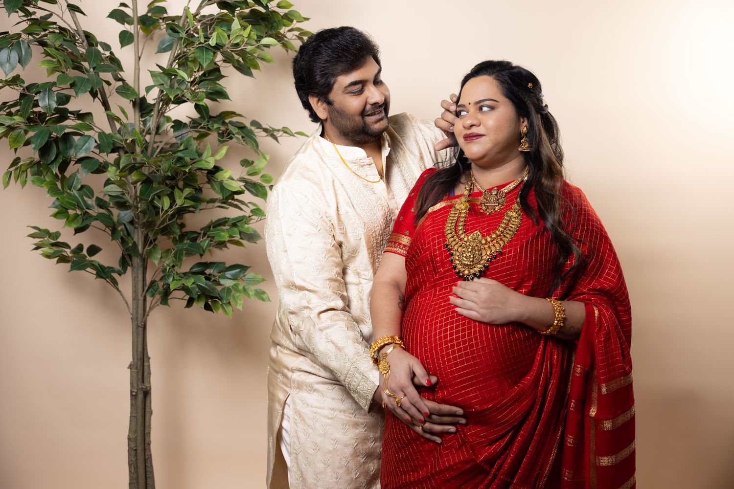 Indian couple in traditional dress, man in white kurta embracing pregnant woman in red saree.