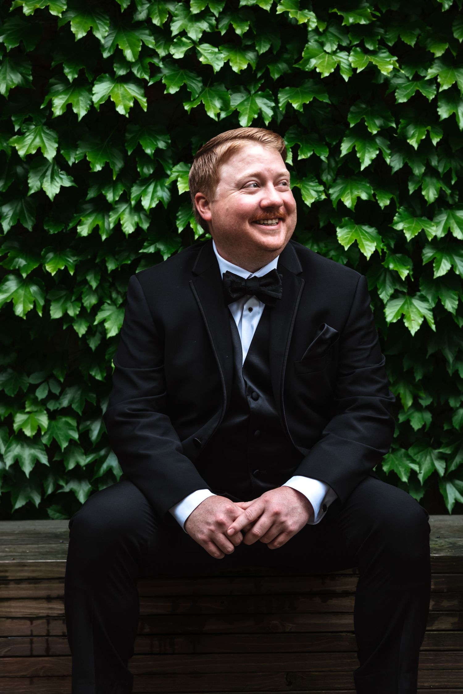 Smiling groom in black tuxedo sits on bench with hands clasped before a dense green ivy wall.