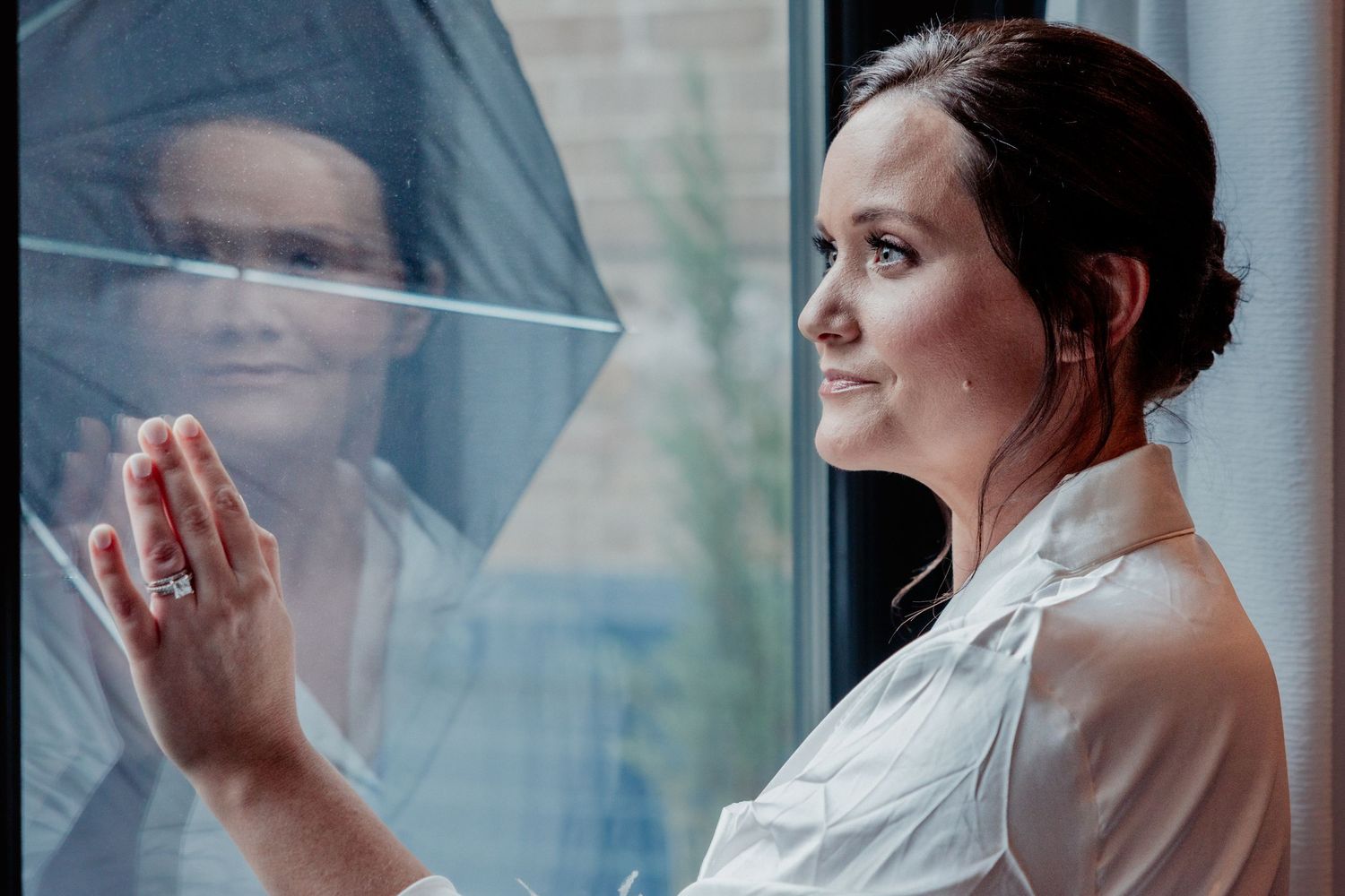 Bride in white robe gazing pensively out window with her reflection in the glass.