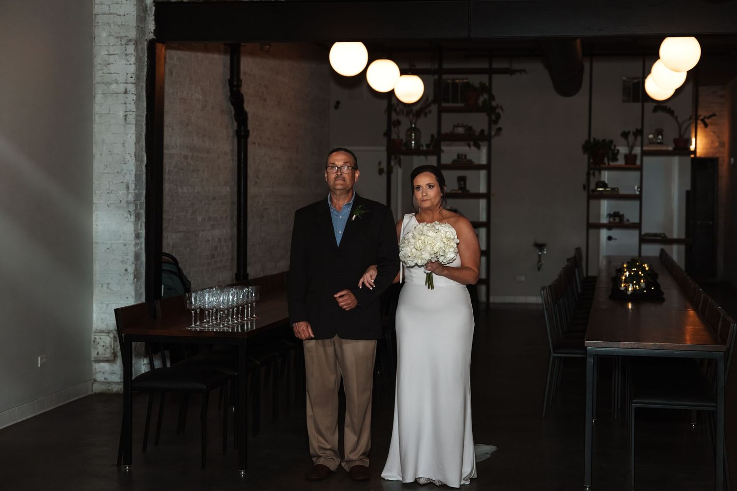 A bride in a white gown walks with her escort through a stylish industrial wedding venue.