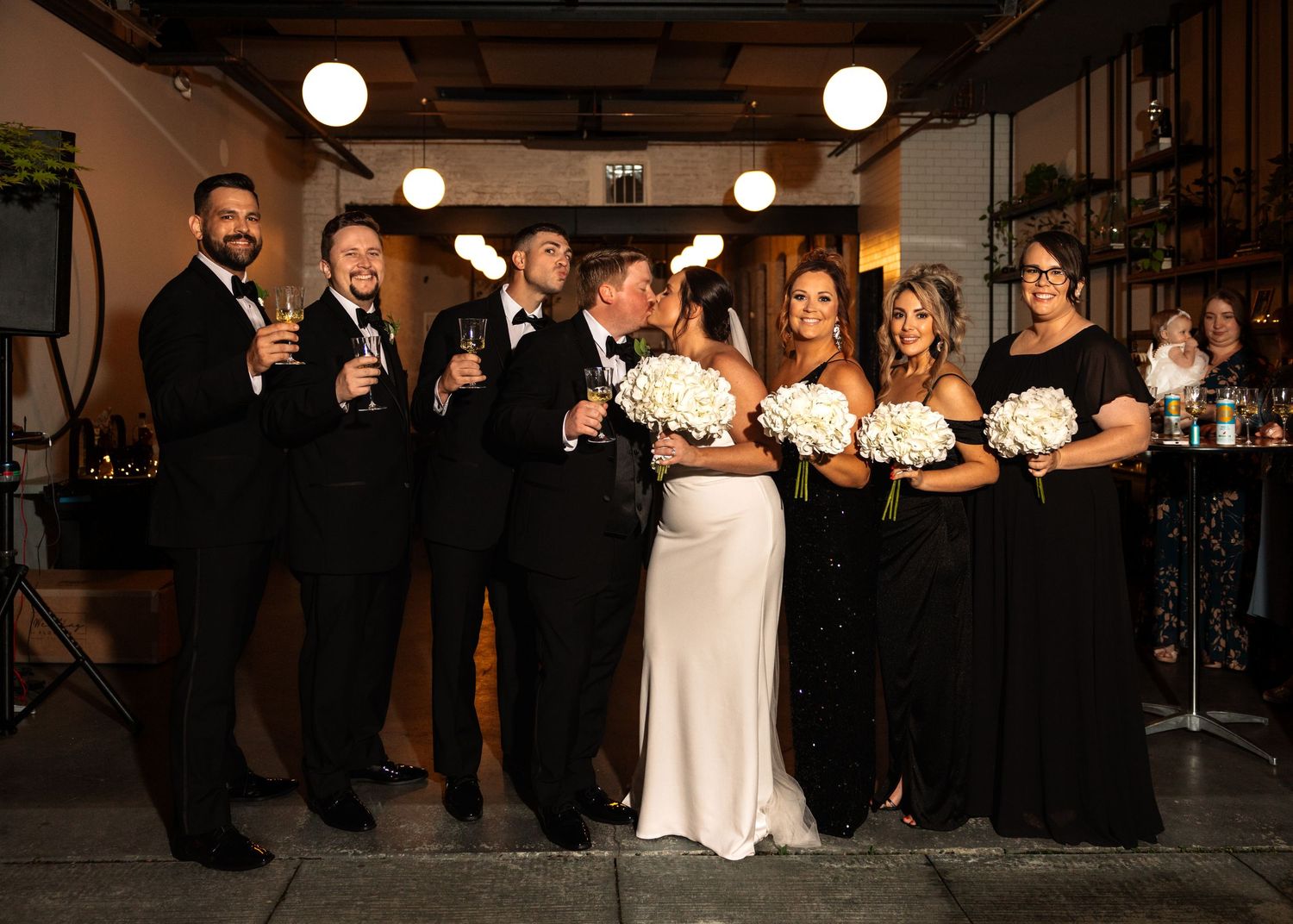 Wedding party in black attire with bride and groom sharing a kiss while holding champagne glasses and white bouquets.