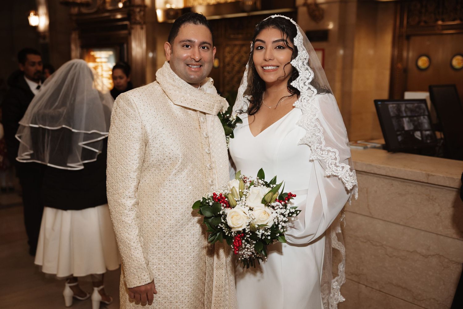Couple posing together at wedding, groom in cream sherwani and bride in white gown with lace veil.