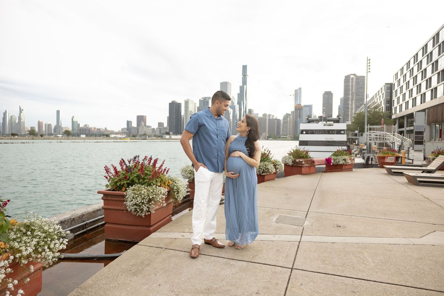 Expectant couple holding hands and smiling on Chicago waterfront dock with city skyline behind them.