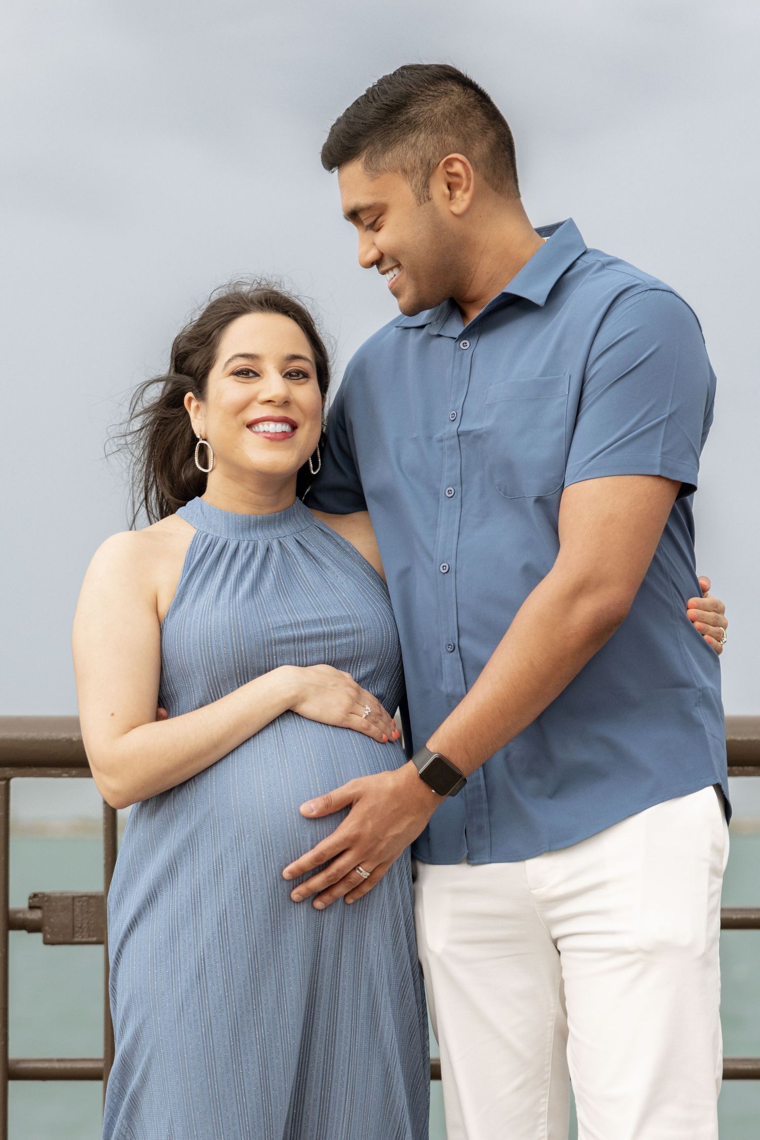 Happy expectant couple in blue outfits tenderly cradling baby bump on an outdoor waterfront location.