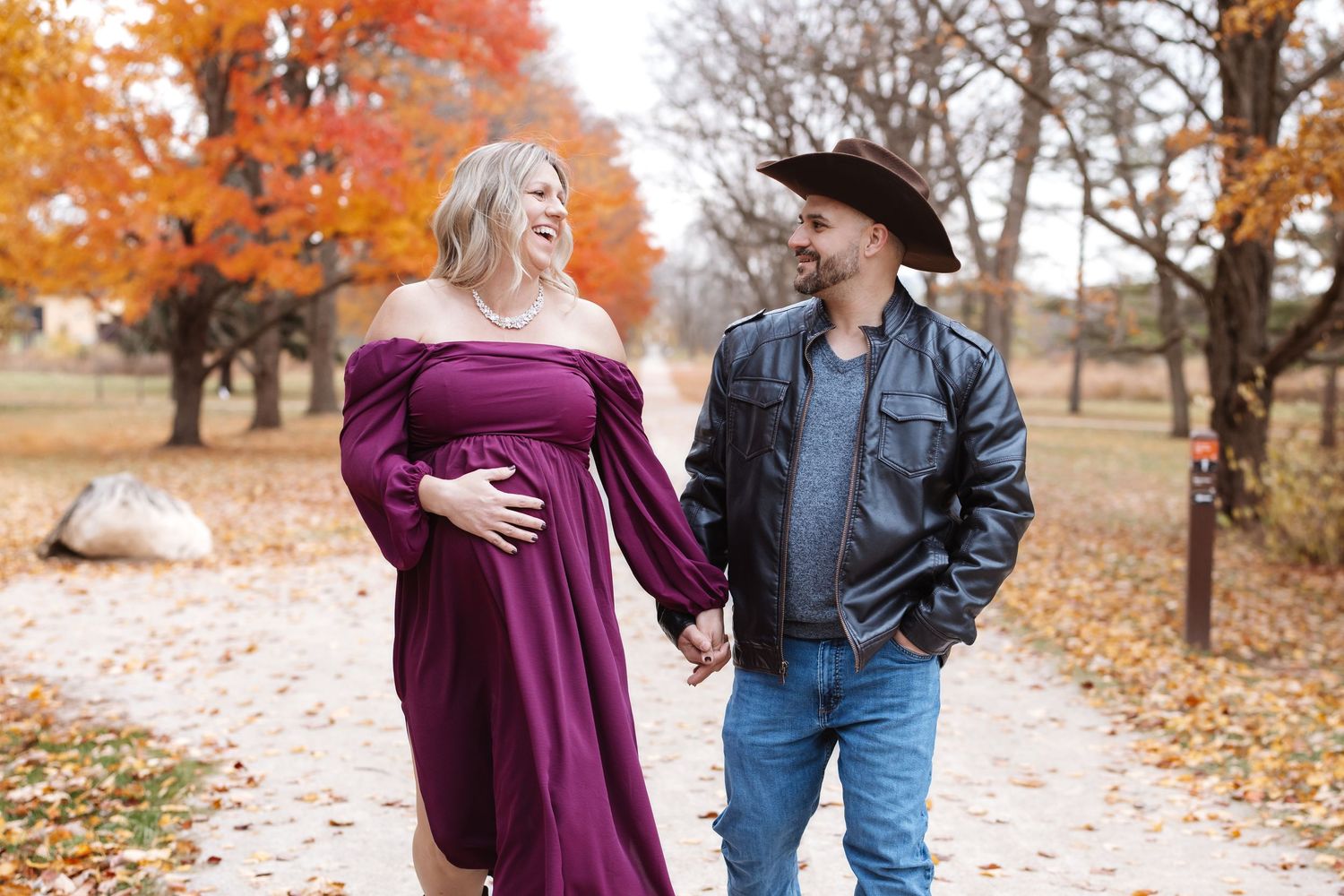 Smiling pregnant couple holding hands while walking on scenic autumn park pathway.