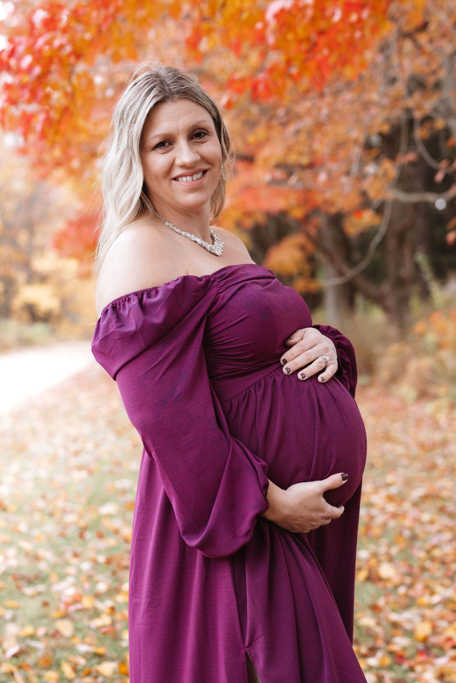 Smiling pregnant woman in elegant purple maternity dress posing outdoors with colorful fall trees.