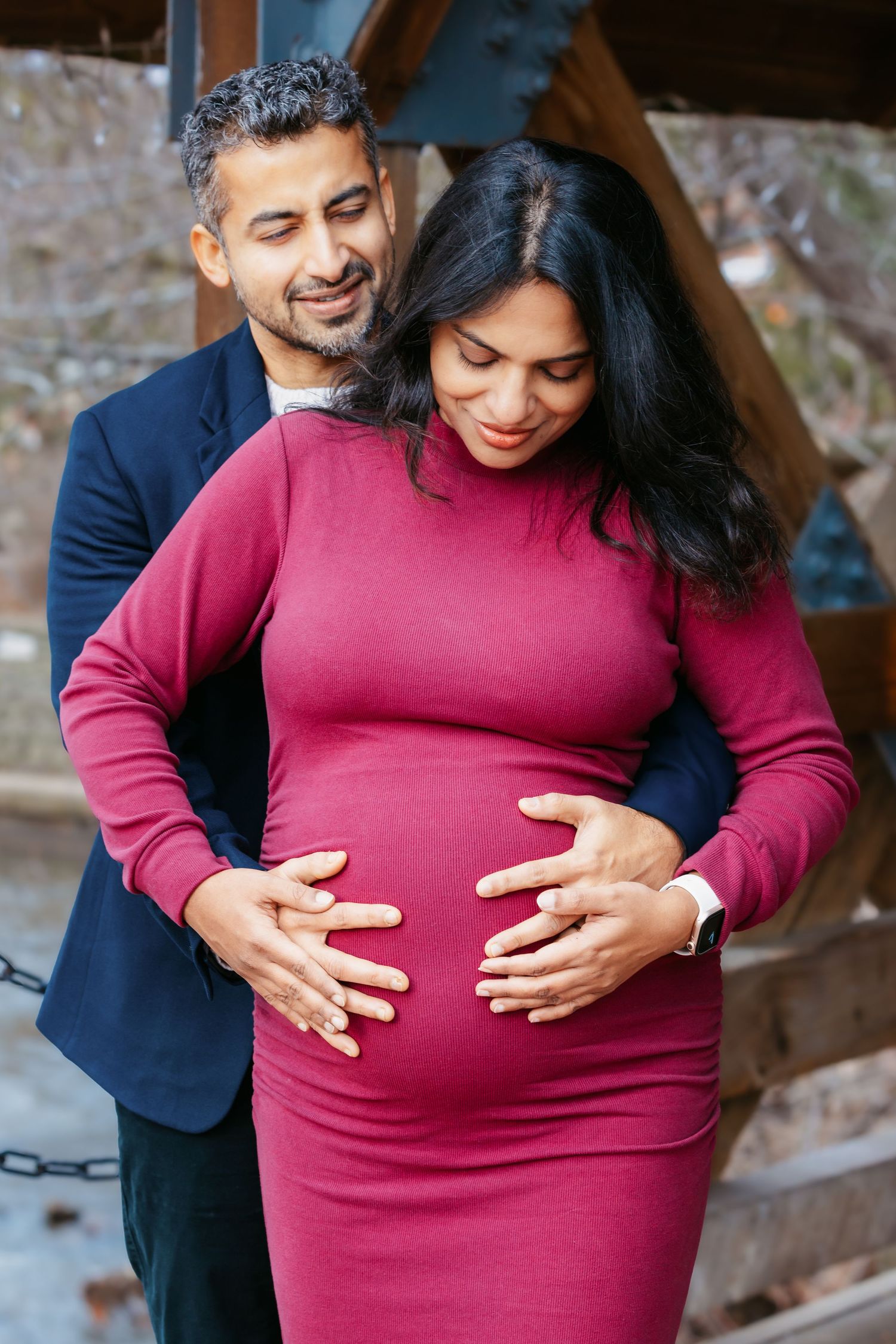 Man tenderly embracing pregnant woman in burgundy dress during outdoor maternity session.