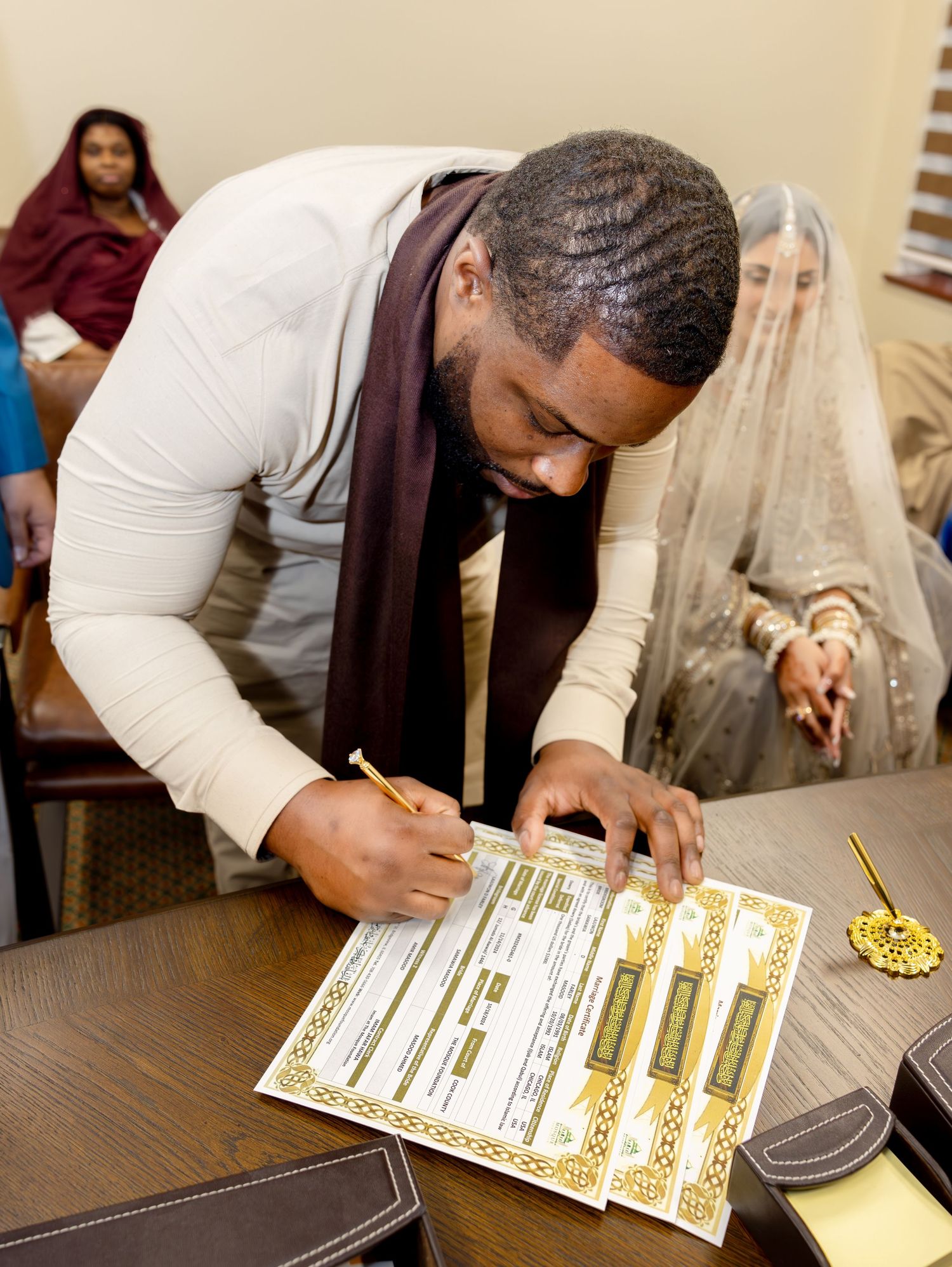 Groom in white outfit signing Islamic Nikkah marriage certificate while bride watches in background.