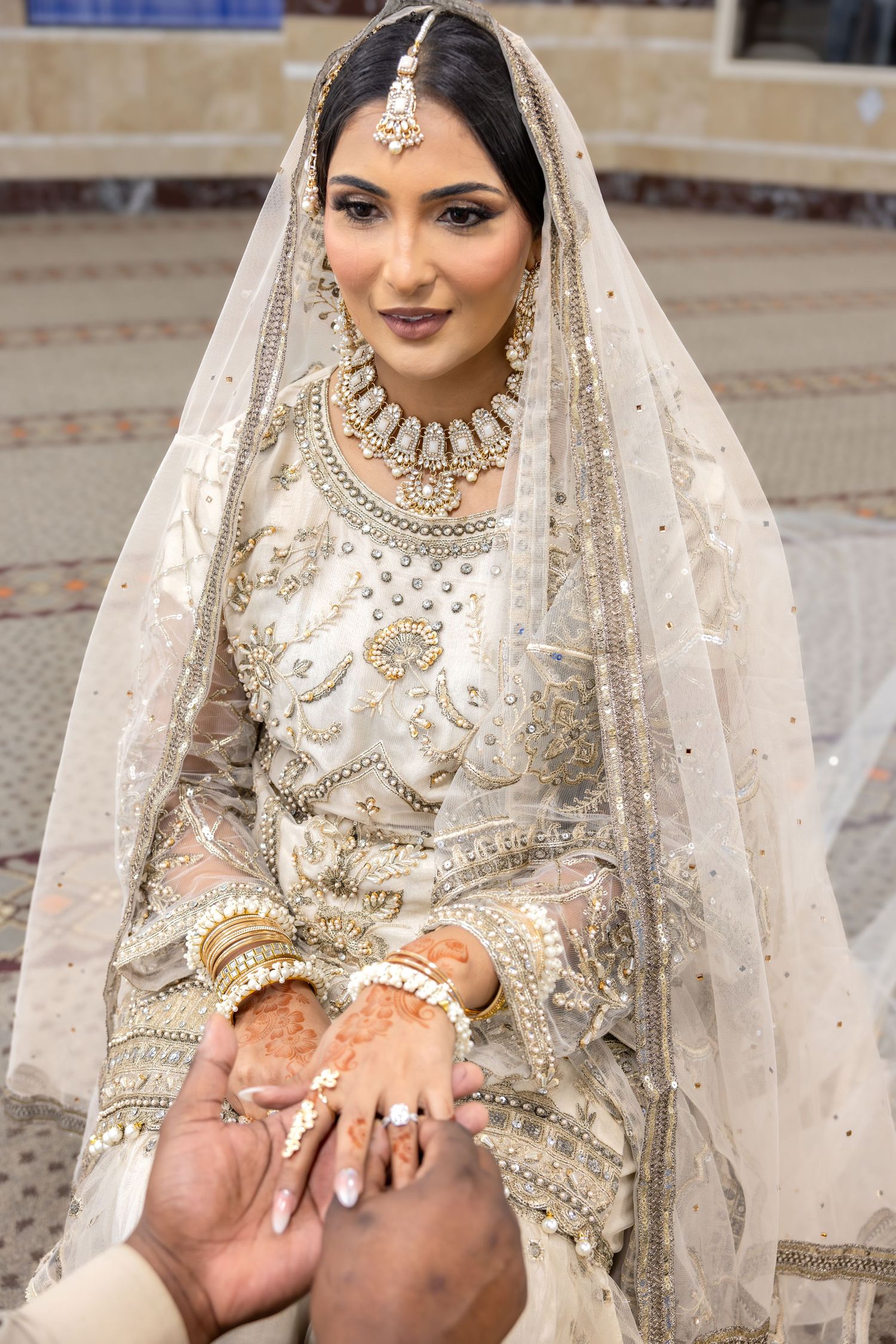 Elegant South Asian bride in white embroidered lehenga with gold jewelry and dupatta holding groom's hands.