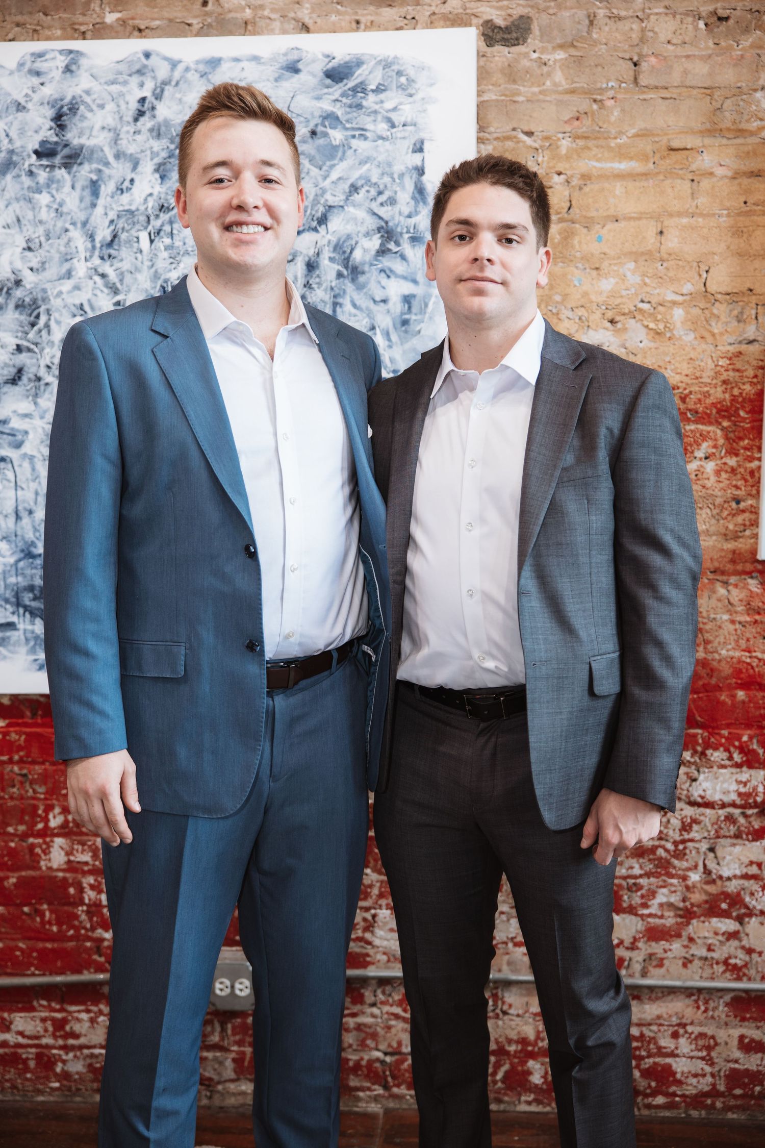 Two smiling men in blue and gray suits pose together in an art gallery with brick walls.