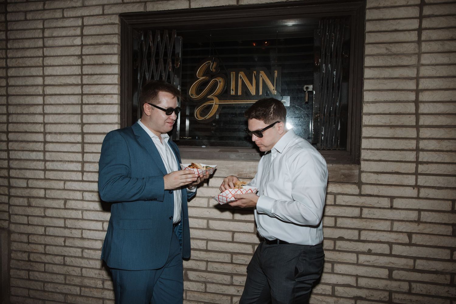 Two men in suits eating food outside an inn at night wearing sunglasses.