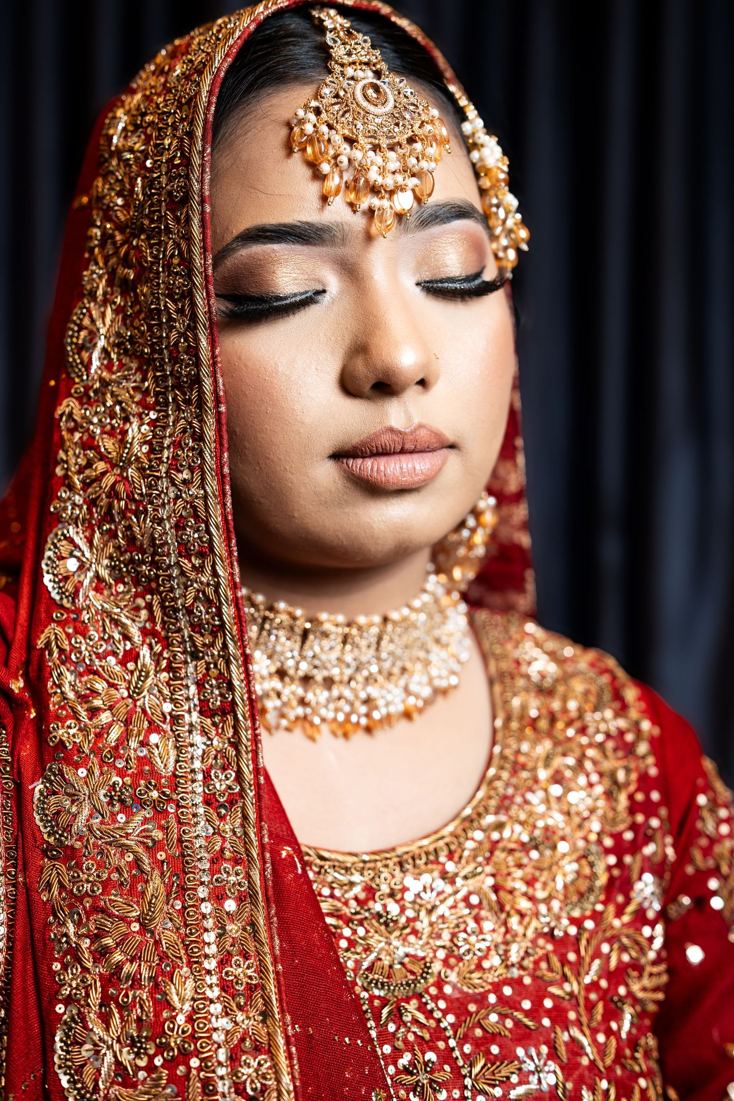Close-up portrait of a bride in red and gold Indian bridal jewelry with eyes closed.
