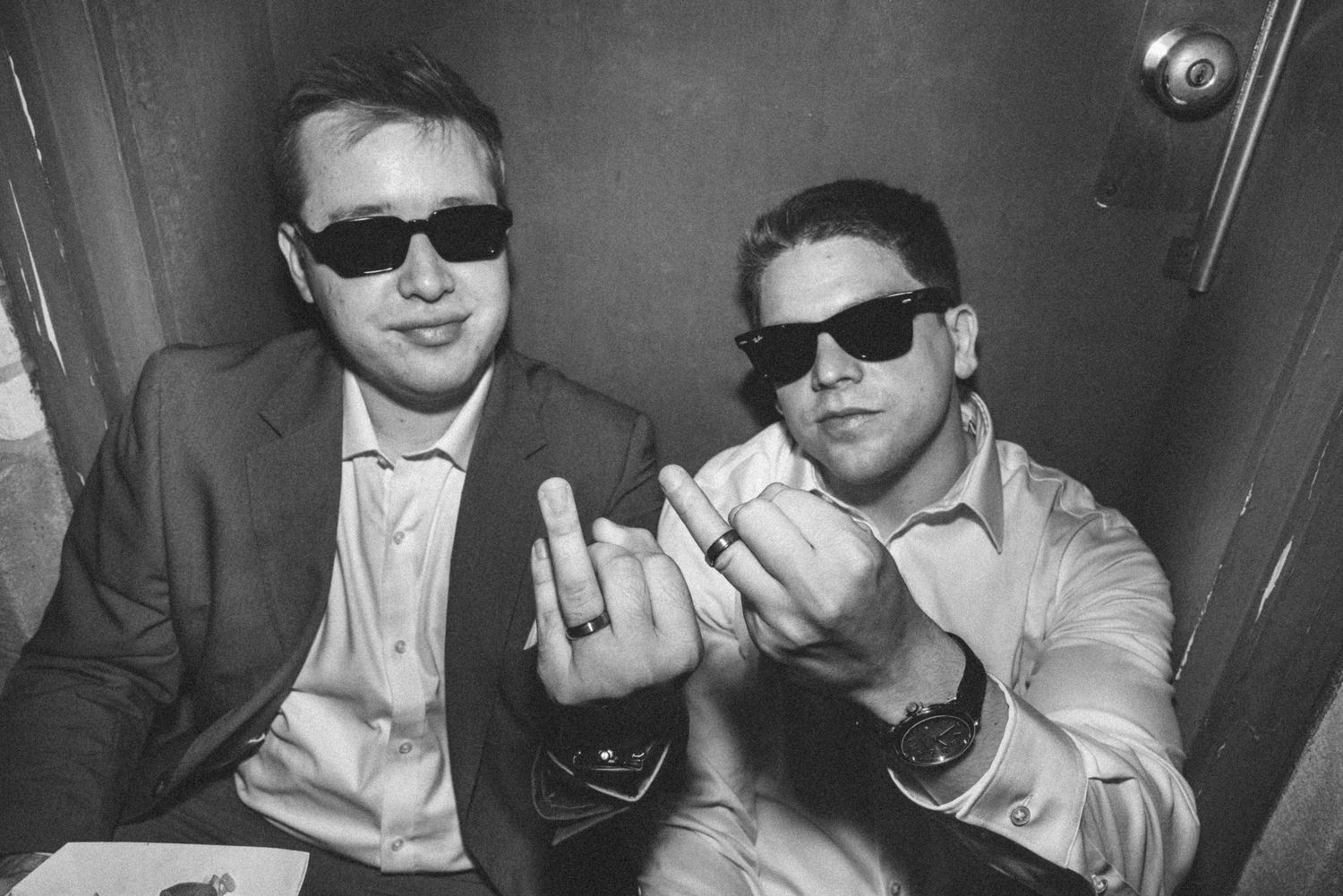 Black and white photo of two men in suits and sunglasses flashing wedding rings in photo booth.