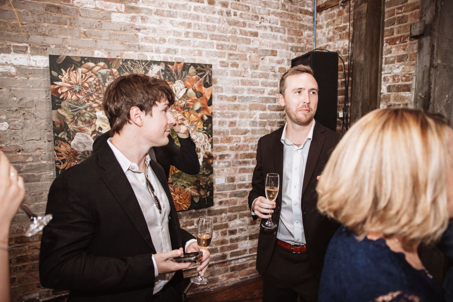 Two men in dark suits conversing near artwork at an upscale brick-walled reception venue.