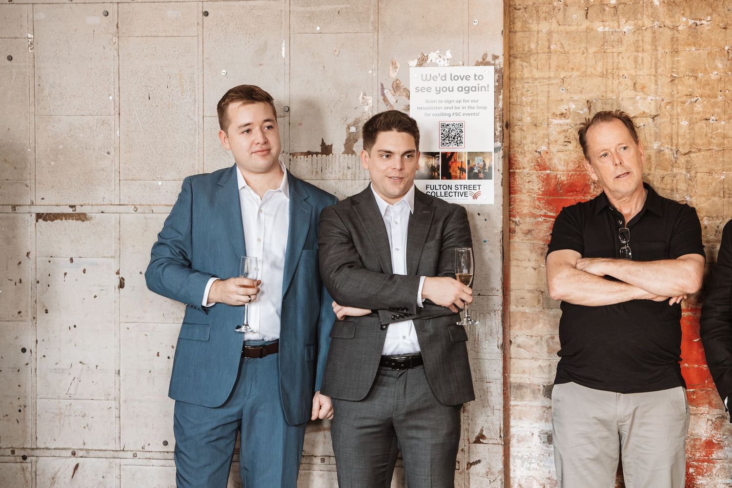 Three men in suits and casual wear stand against concrete wall at art gallery event.