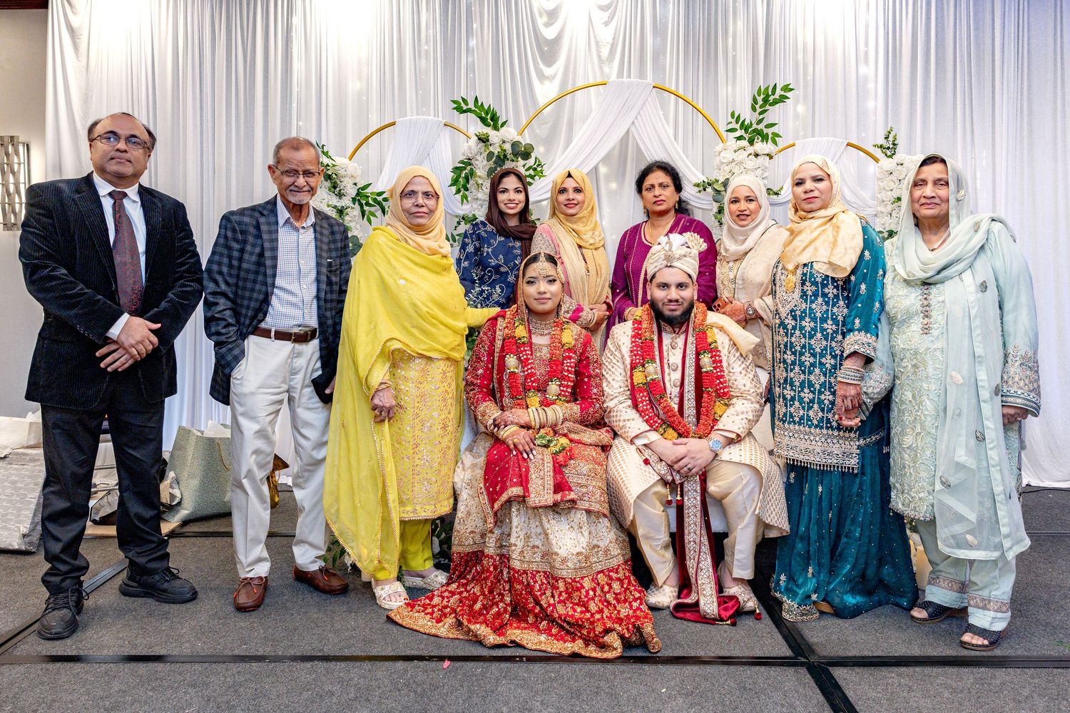 Wedding party group photo with South Asian bride and groom in traditional attire before floral arch.