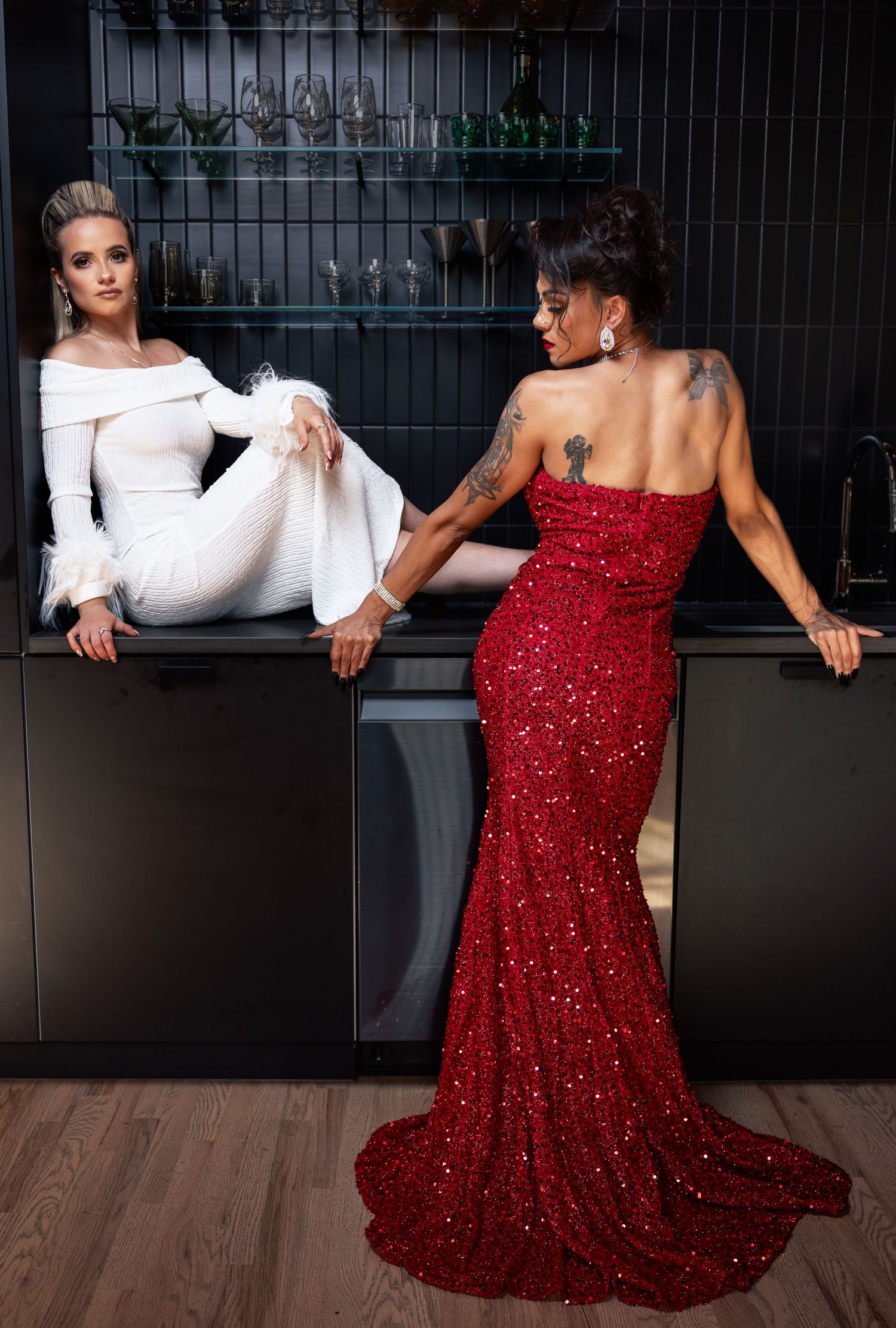 Two women in white and red sequin gowns posing in dark elegant kitchen setting.