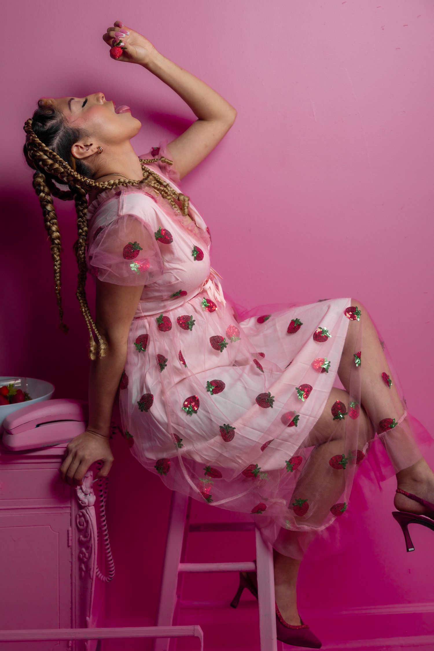 Woman in pink strawberry tulle dress reclines on stool dropping strawberry into mouth.