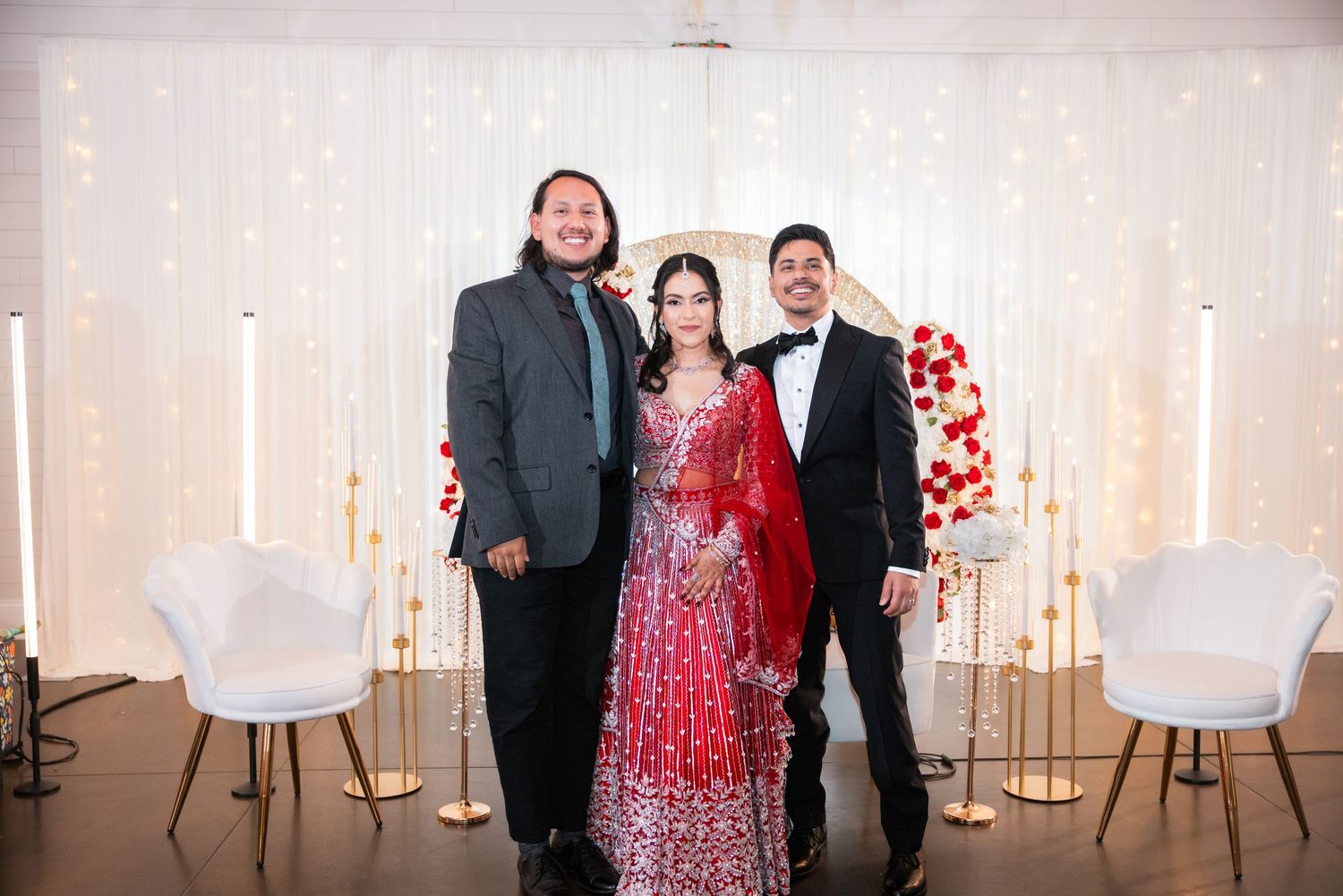 Bride in red lehenga and groom in black tuxedo smile with a guest at elegant wedding venue.