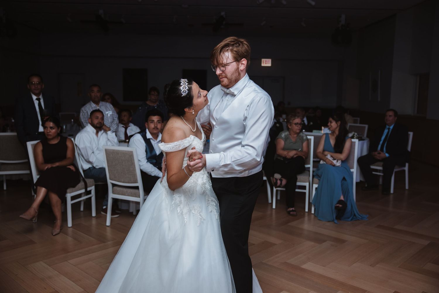 Bride and groom sharing their first dance in white ballroom while wedding guests watch from surrounding tables.