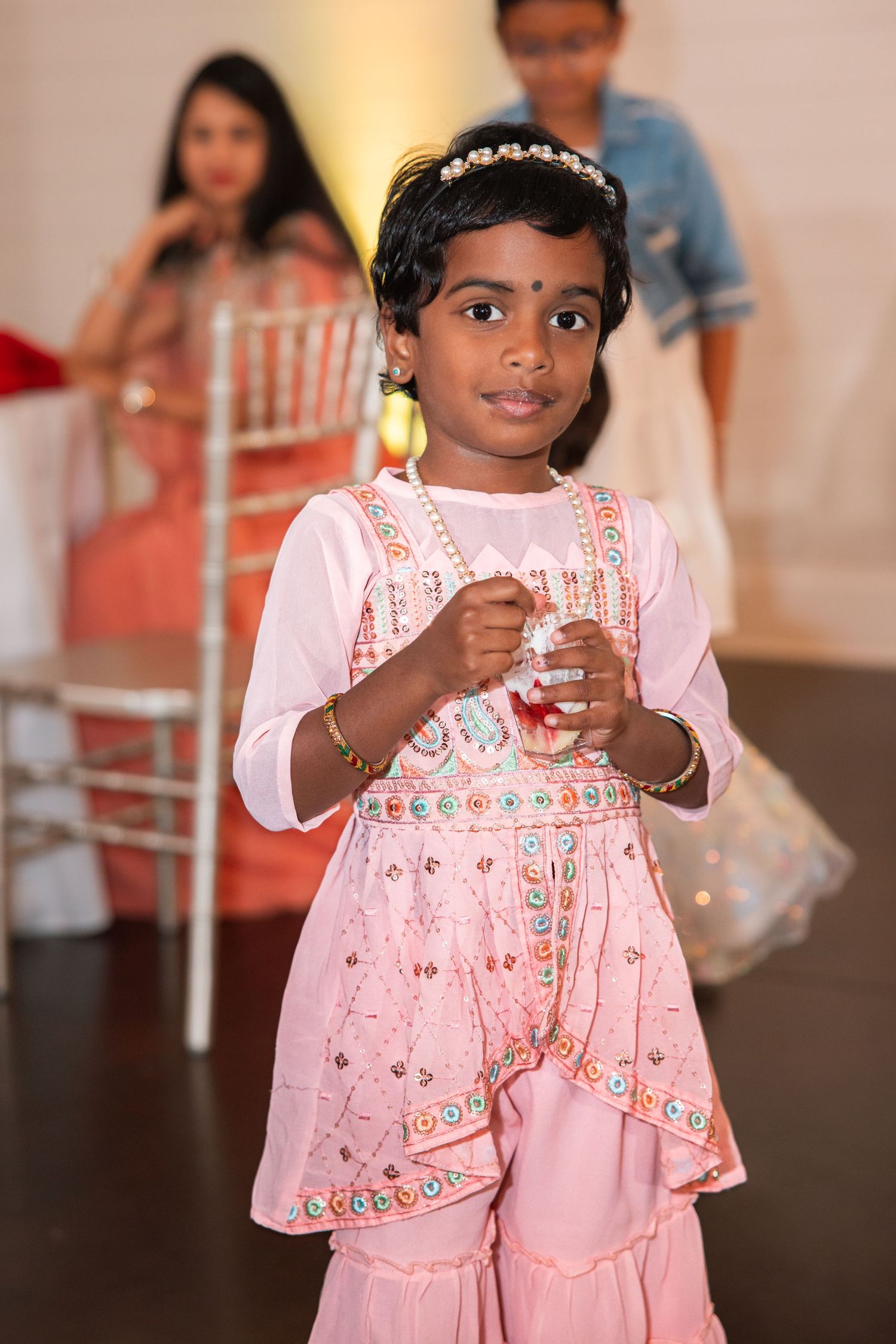 Young Indian girl in pink embroidered salwar kameez with floral headband at a festive celebration event.