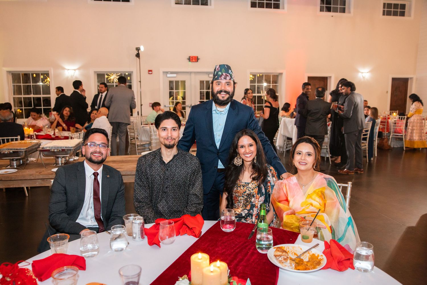 Five people posing together at an elegant South Asian wedding reception dinner in a decorated banquet hall.
