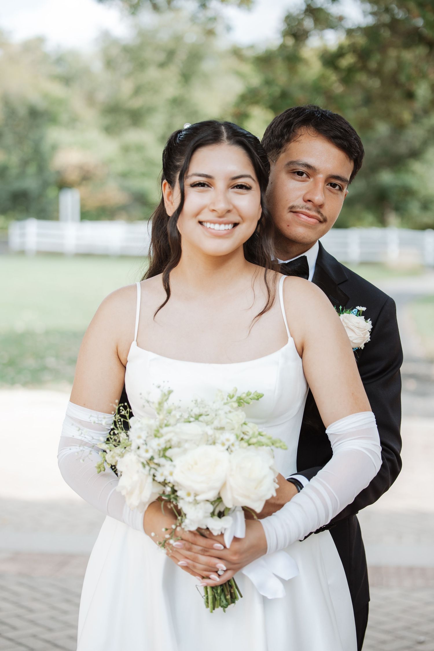 Smiling bride in white dress and gloves holding white rose bouquet with groom in tuxedo.