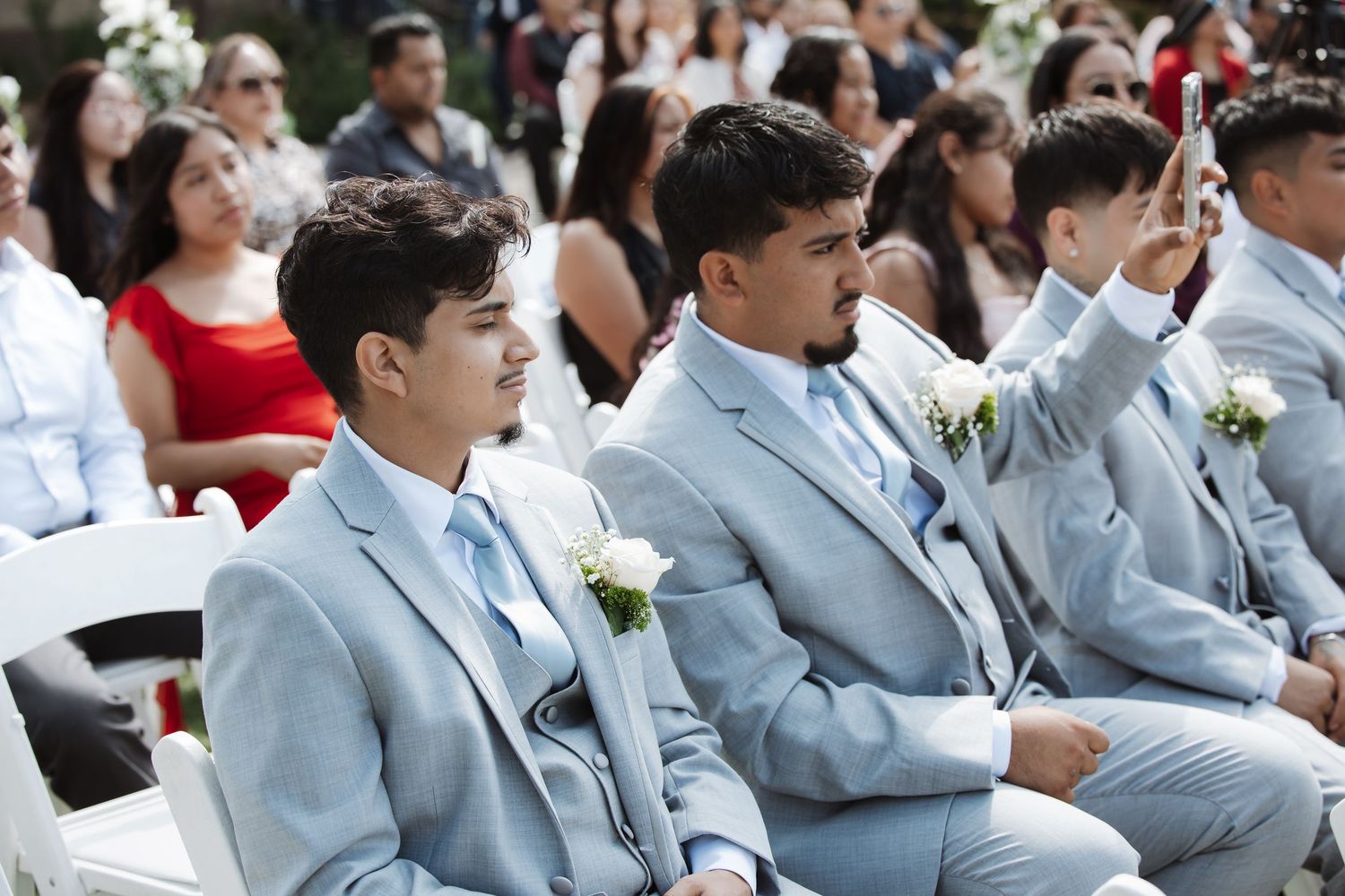 Groomsmen in light blue suits with white boutonnieres seated at outdoor wedding ceremony.