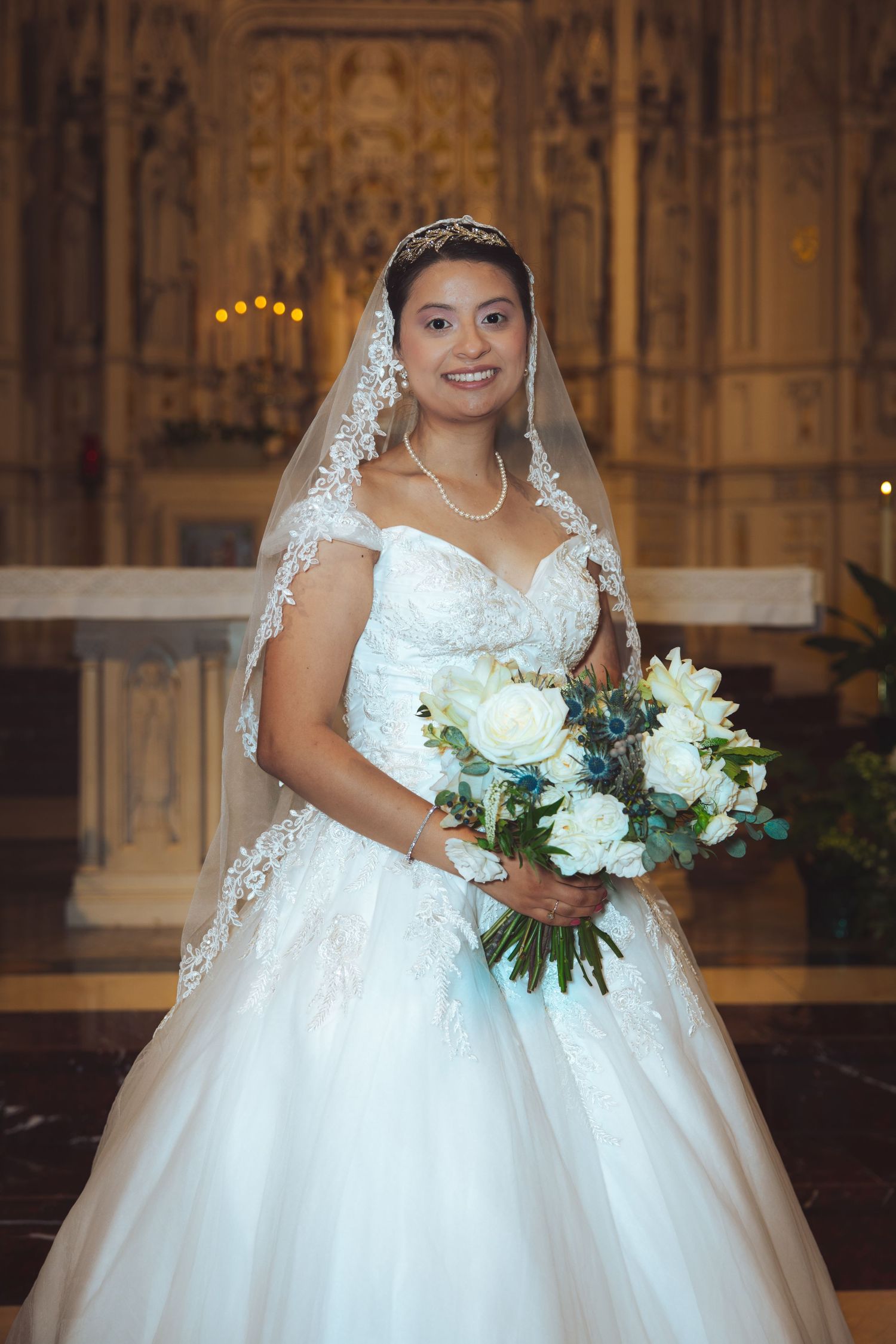 Smiling bride in white lace wedding dress holding bouquet at church altar.