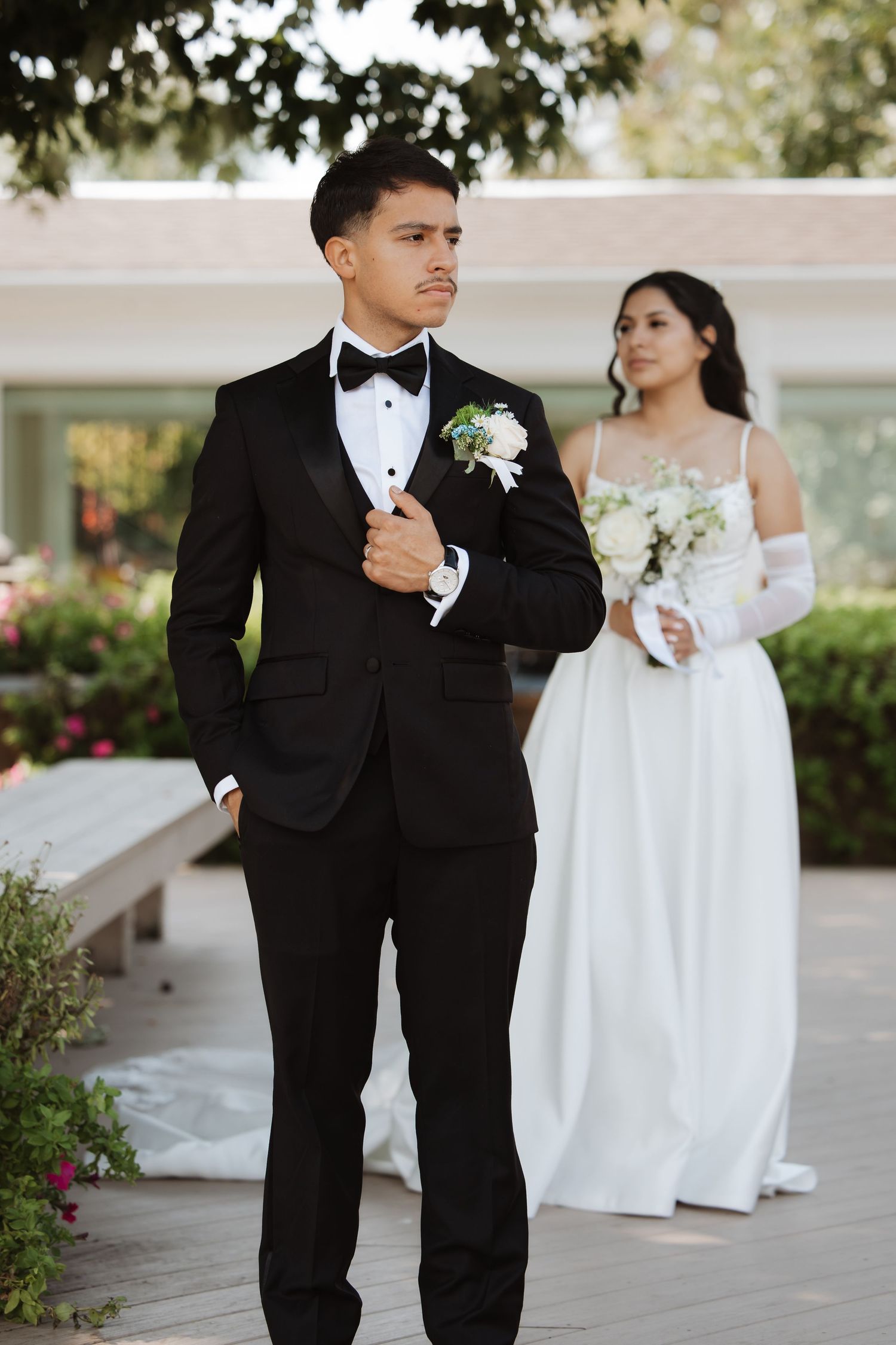 Groom in black suit adjusts jacket while bride in white gown with gloves holds bouquet in background.