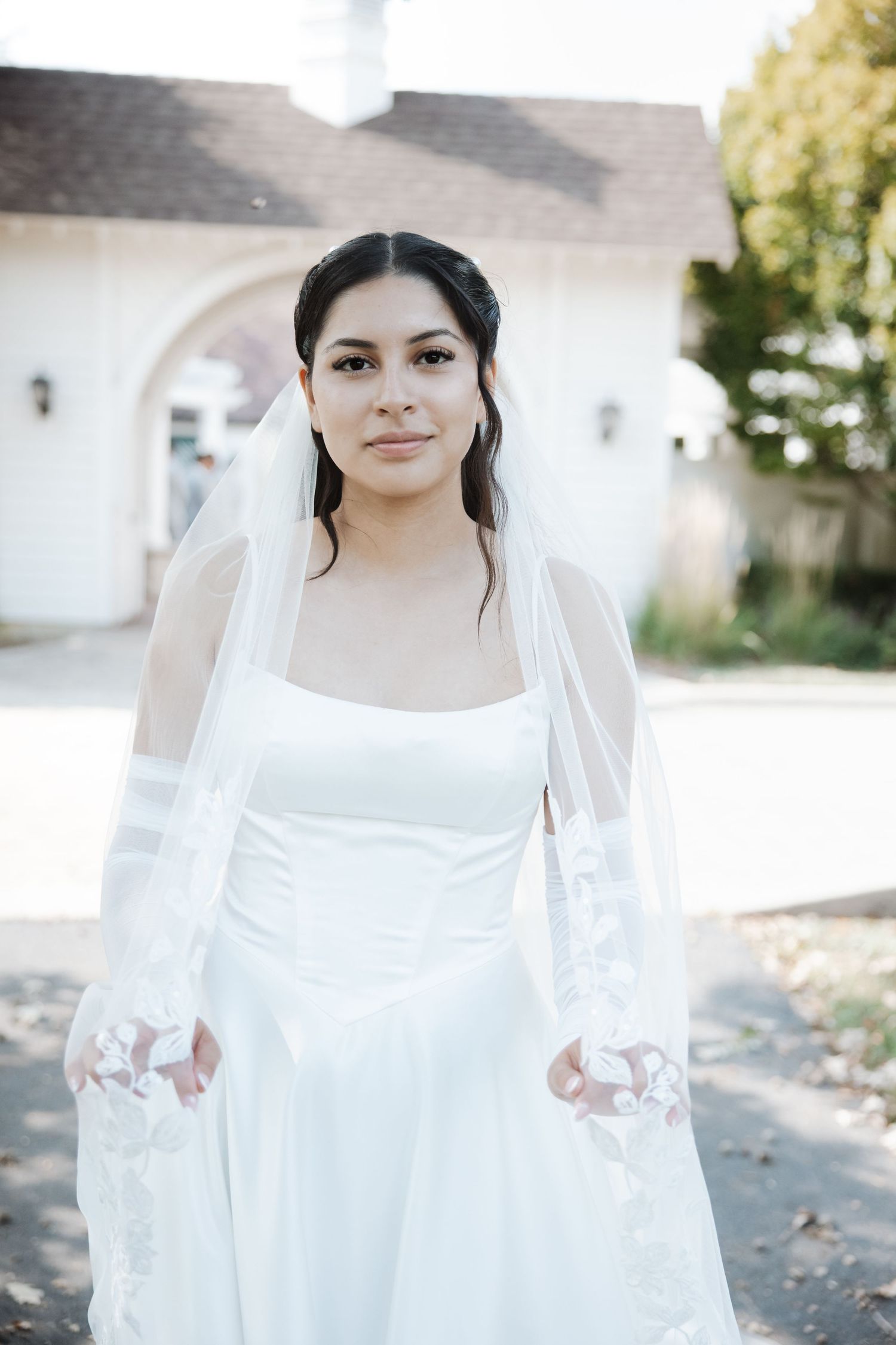 Elegant bride in white square-neck gown and lace veil posing outside white chapel.