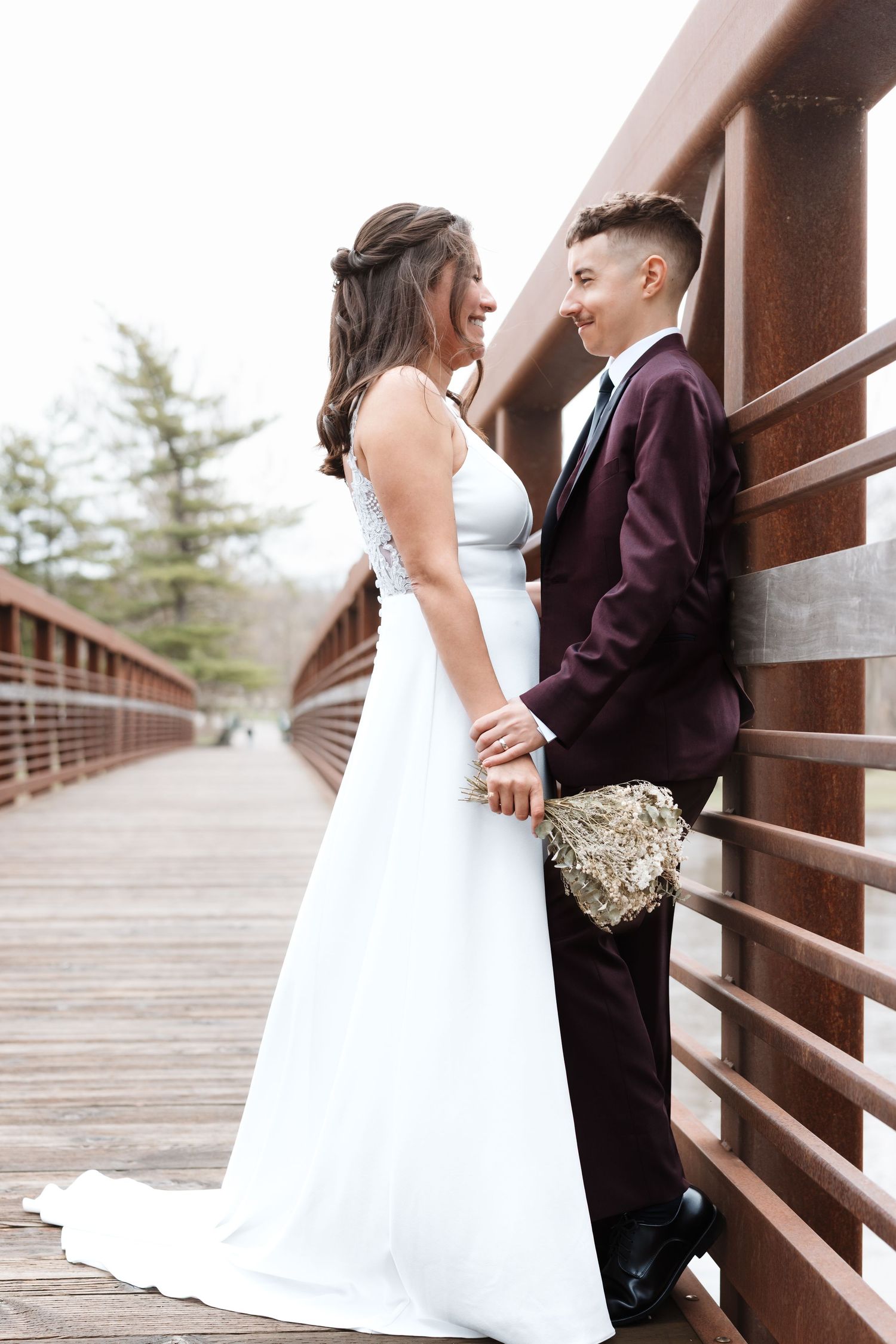 A bride in white gown and partner in burgundy suit hold hands smiling on a wooden bridge.