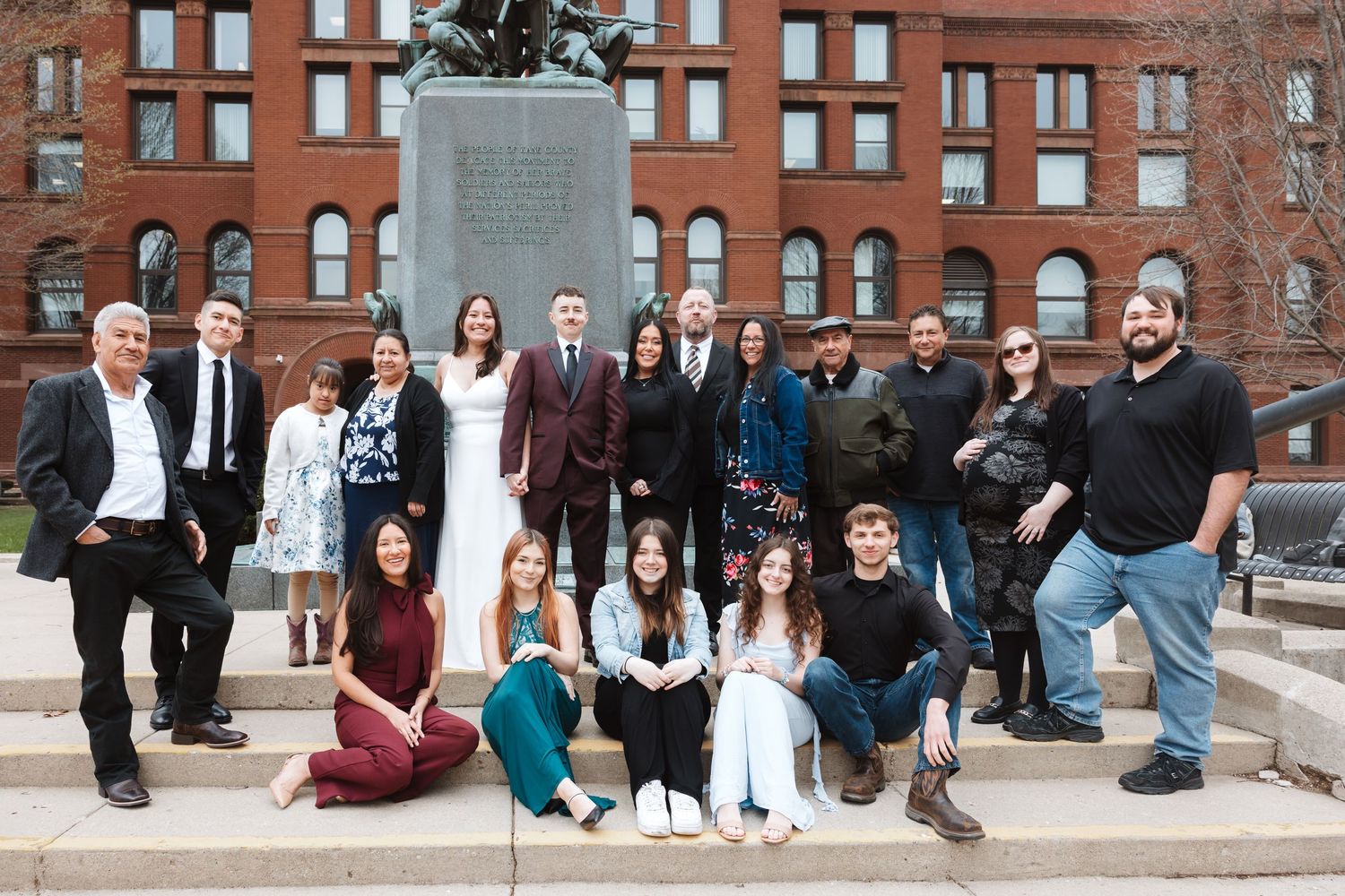 Large group of people posing outdoors near a statue and brick building for a formal occasion.