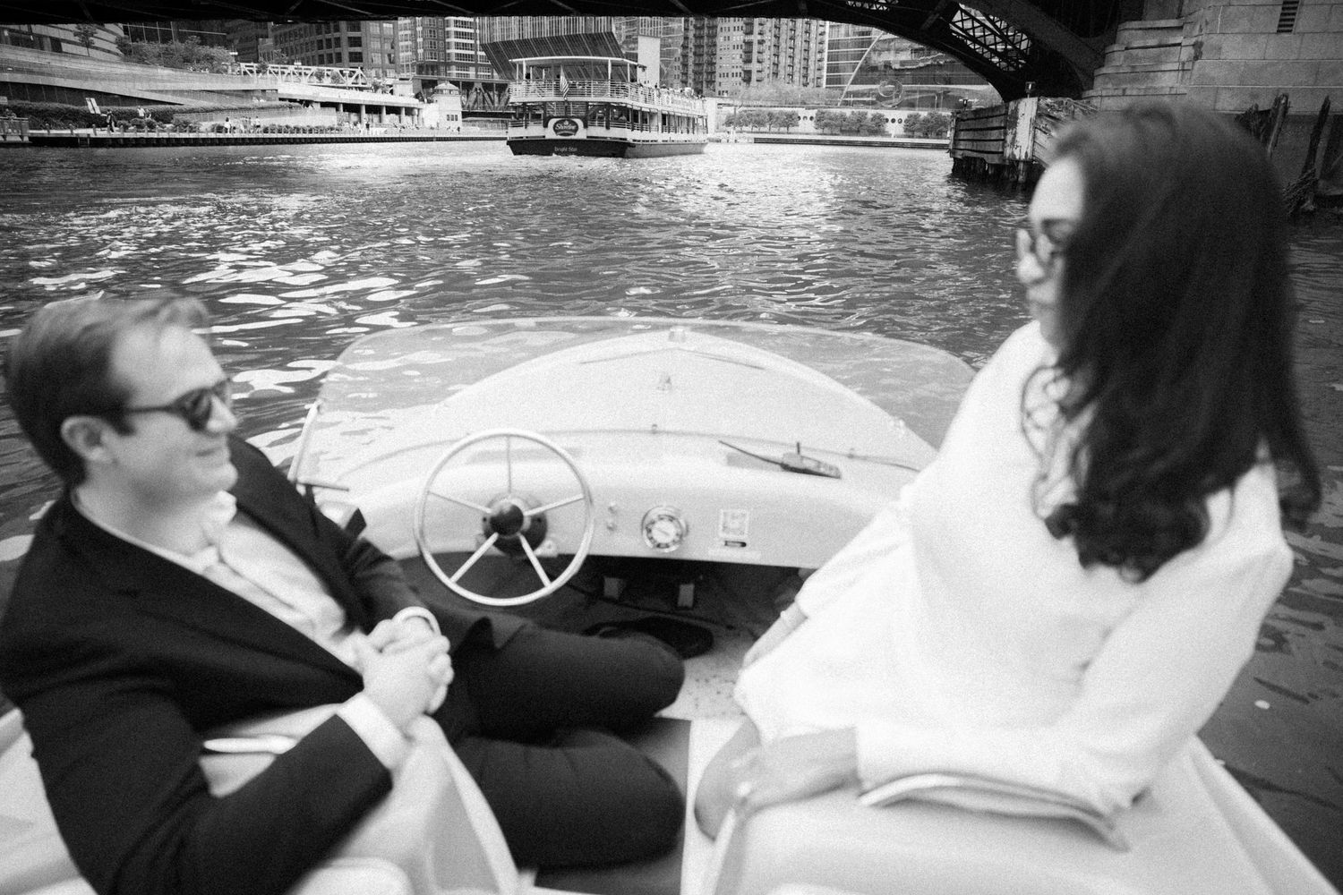 Black and white photo of a couple riding a vintage boat on a city waterway during an engagement session.