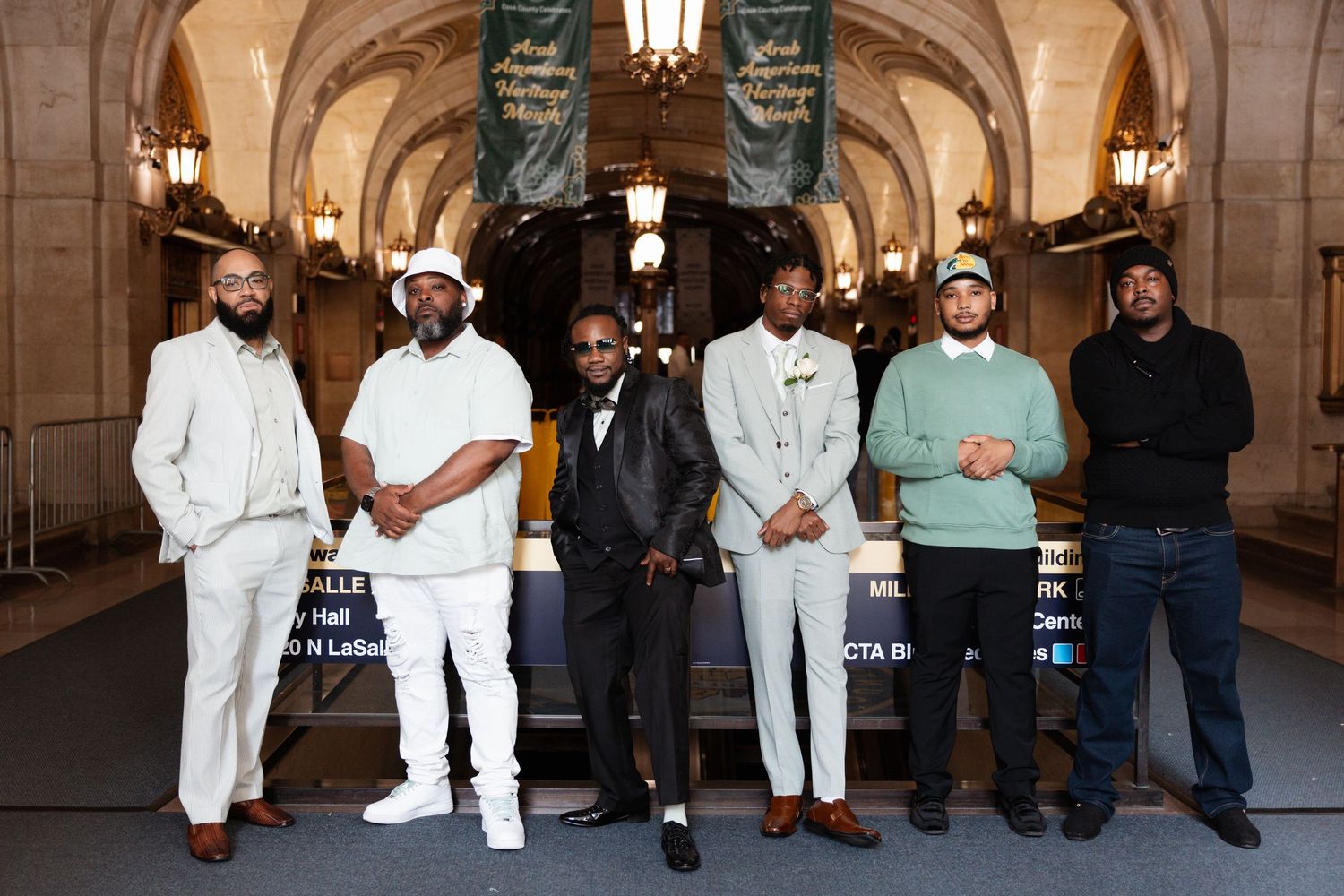 Six men in formal attire pose together inside an elegant building with decorative lighting.