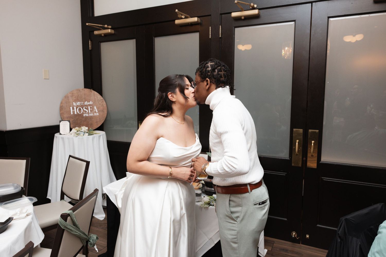 Bride and groom share a kiss near personalized wooden wedding sign at reception.