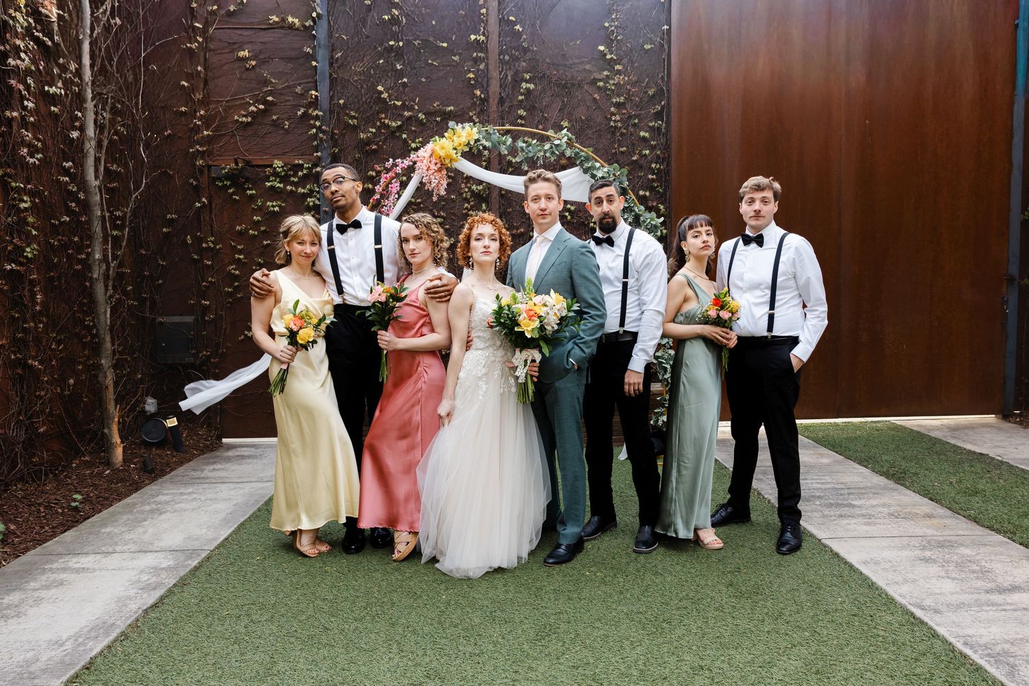 Wedding party portrait with bride in white gown, groom in teal suit, bridesmaids and groomsmen outdoors.